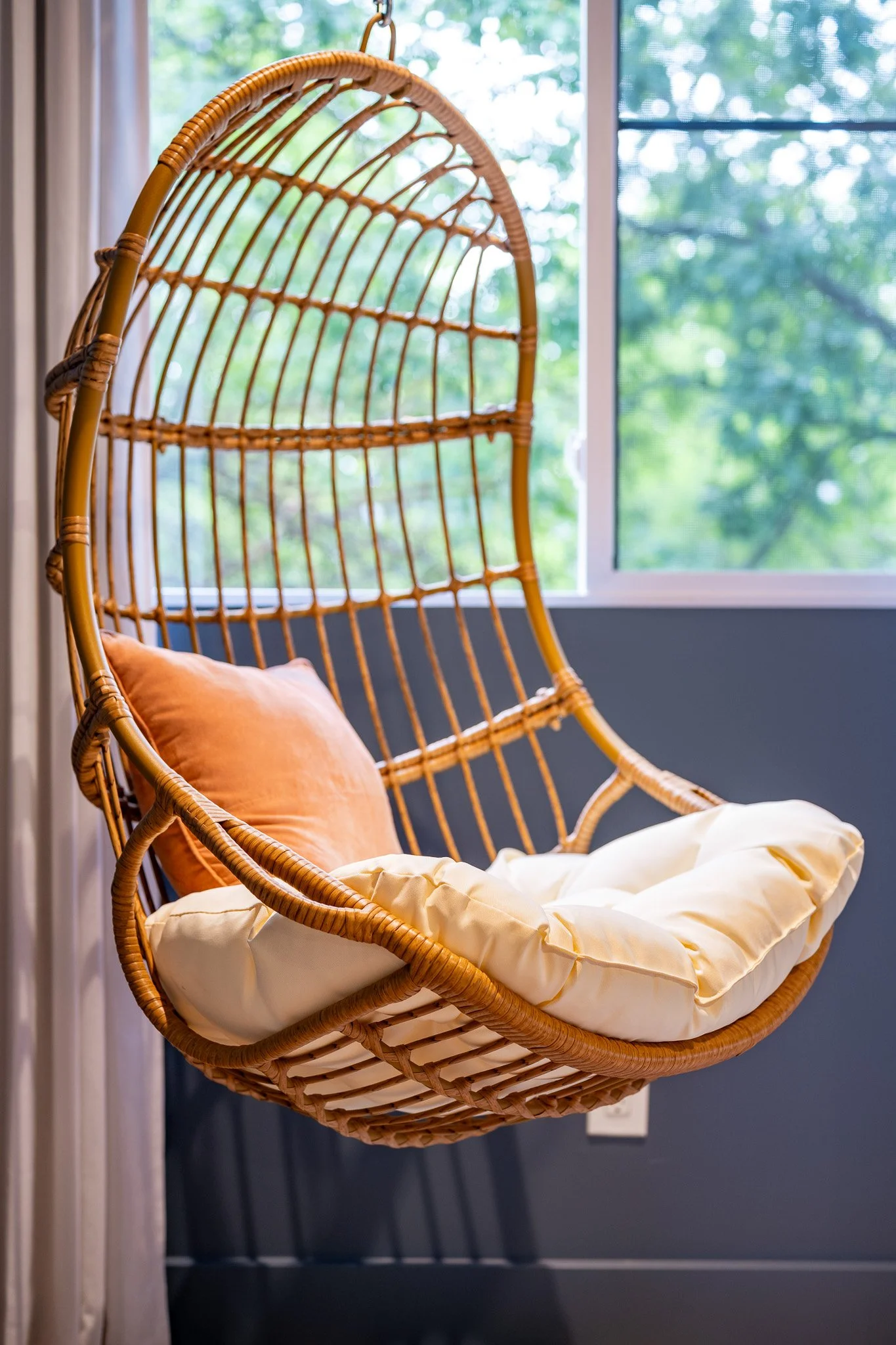 A hanging rattan chair with a peach-colored cushion and a beige blanket, hanging near a window with a view of green trees outside.