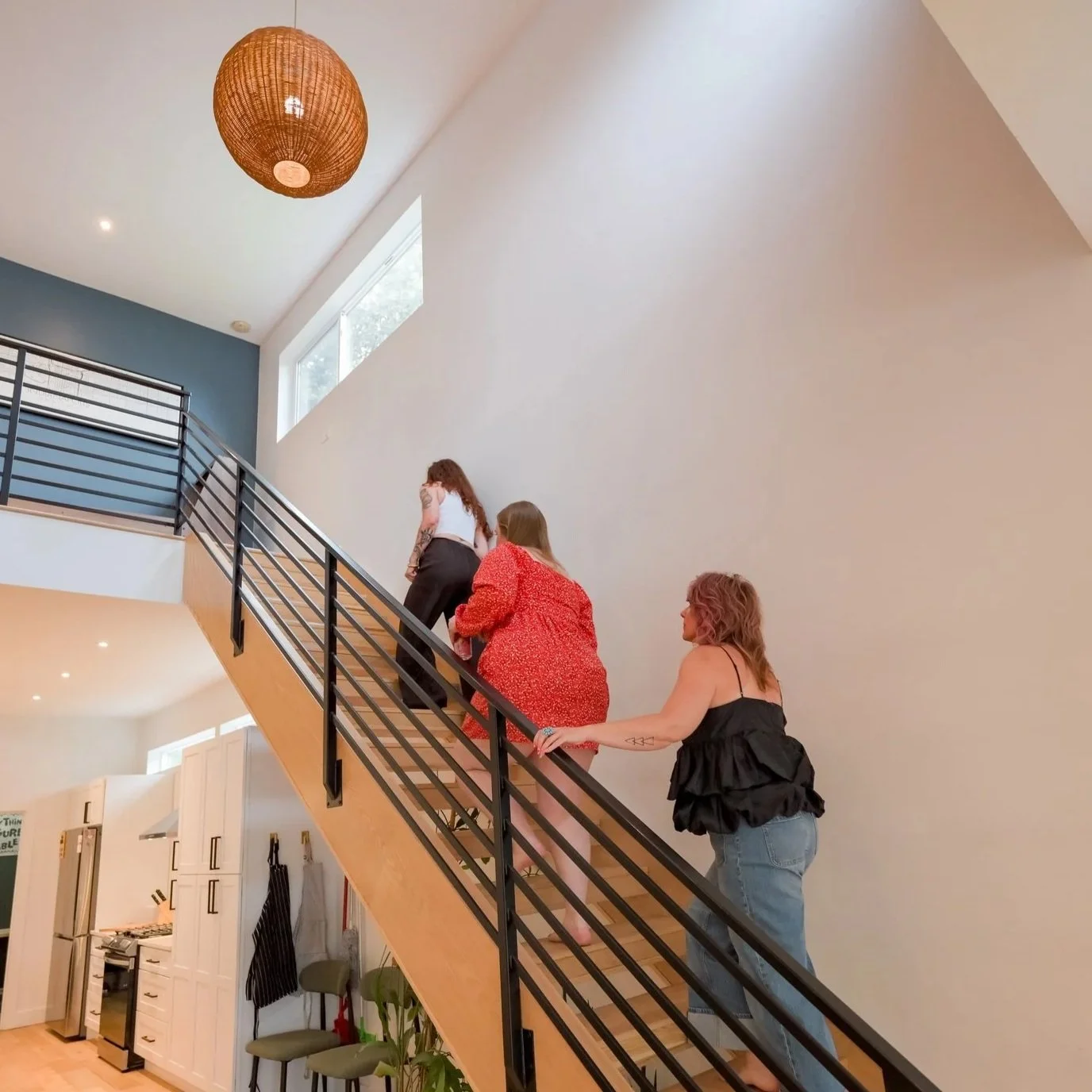 Three women ascending a staircase in a modern home interior, with a large window and a hanging wicker light fixture visible.