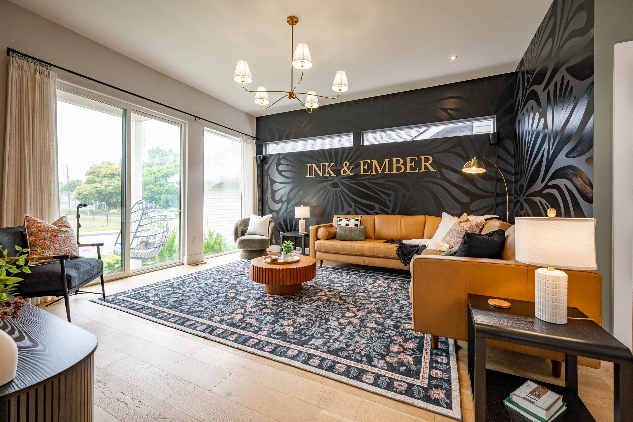 Living room with large sliding glass doors, patterned rug, beige and black sofa, gray armchair, hanging chandelier, and black accent wall with gold lettering that reads 'INK & EMBER'.