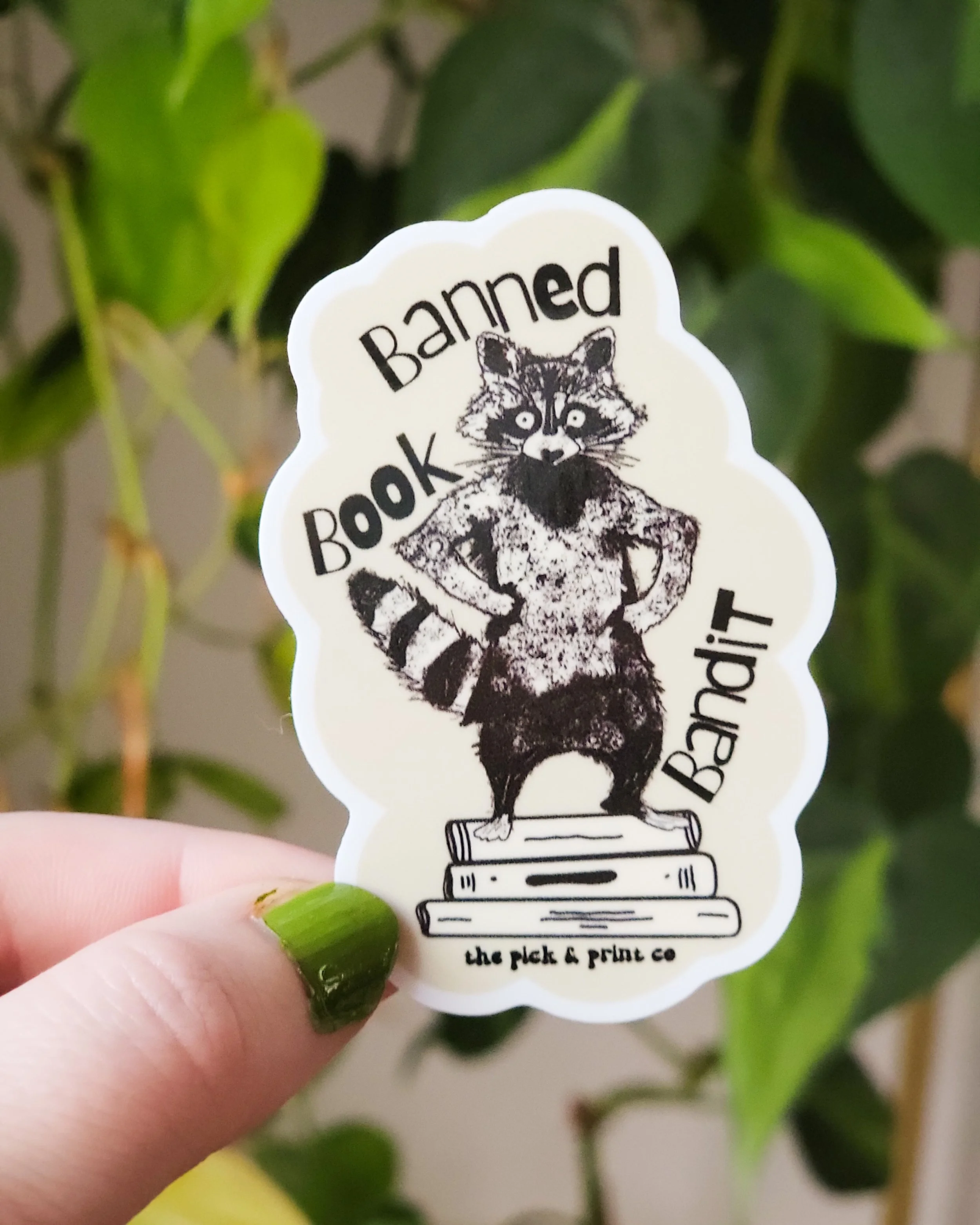 Banned Book Bandit text with a drawing of a raccoon standing on a stack of books, cream background, 3in sticker