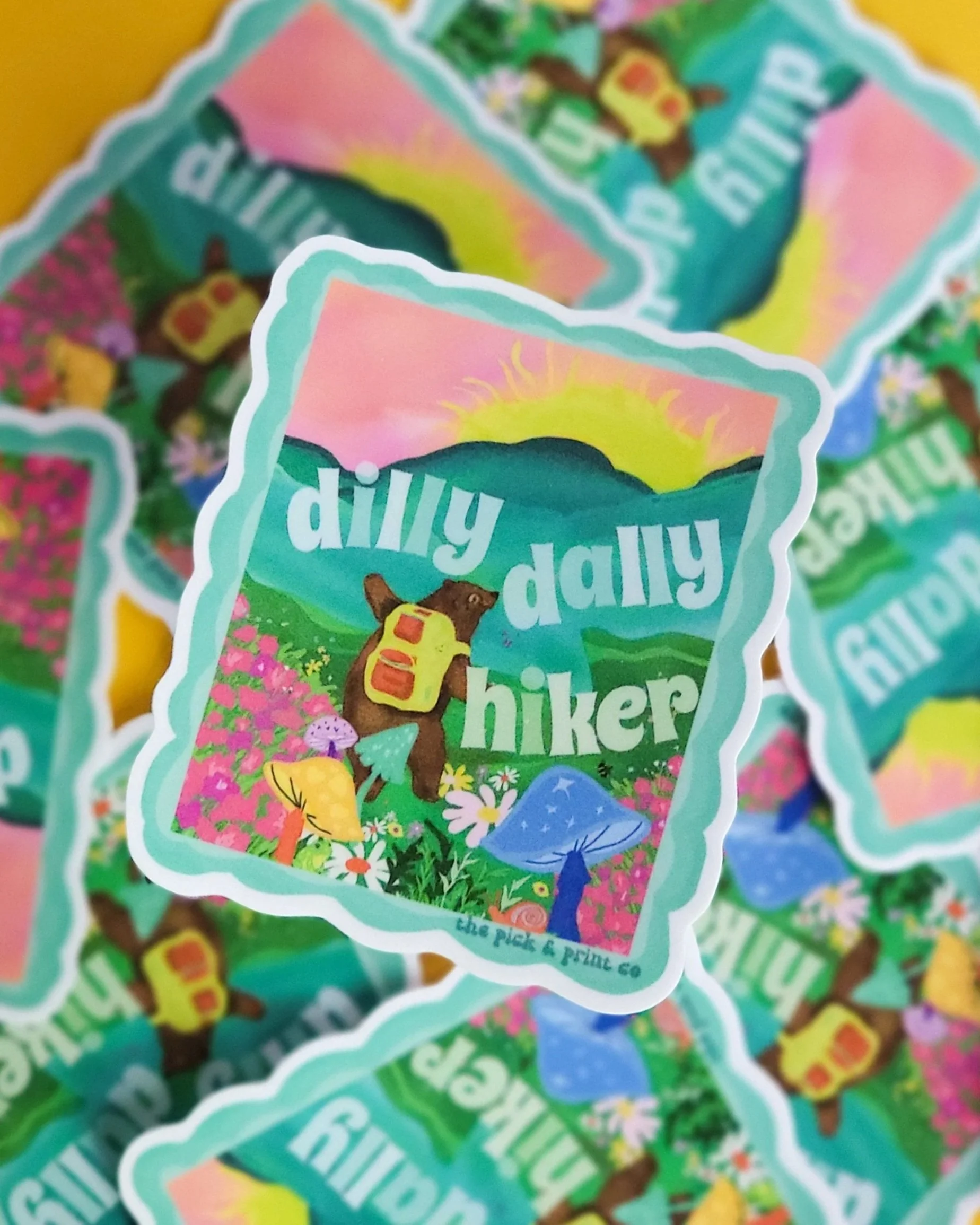Image is of a pile of stickers that have a landscape design on them with a bear hiking and frolicking through wild fowers with the text dilly dally hiker written around the landscape