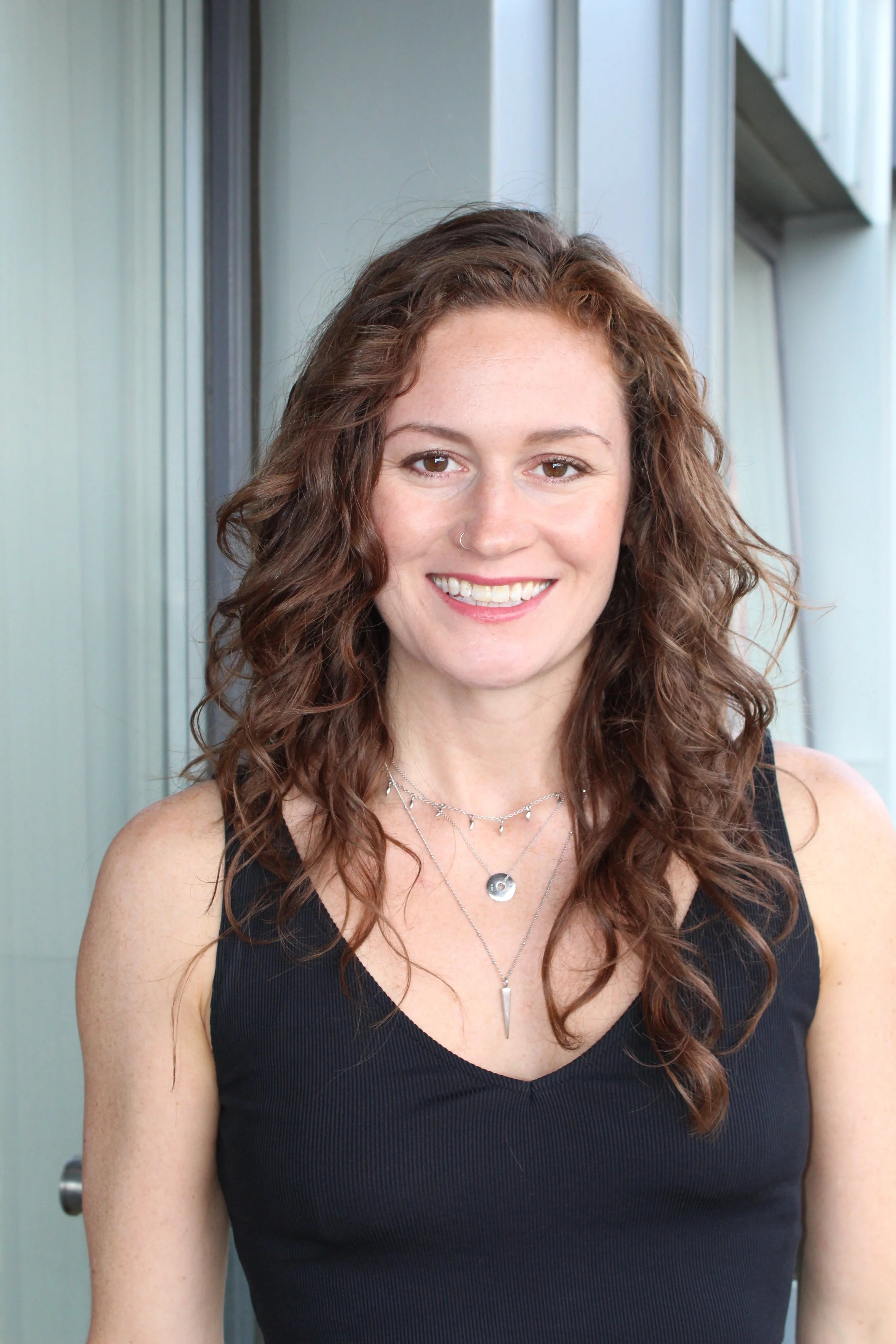 A young woman with long, curly brown hair and a nose piercing smiling at the camera, wearing a black tank top and layered necklaces, standing in front of a modern building with glass panels.