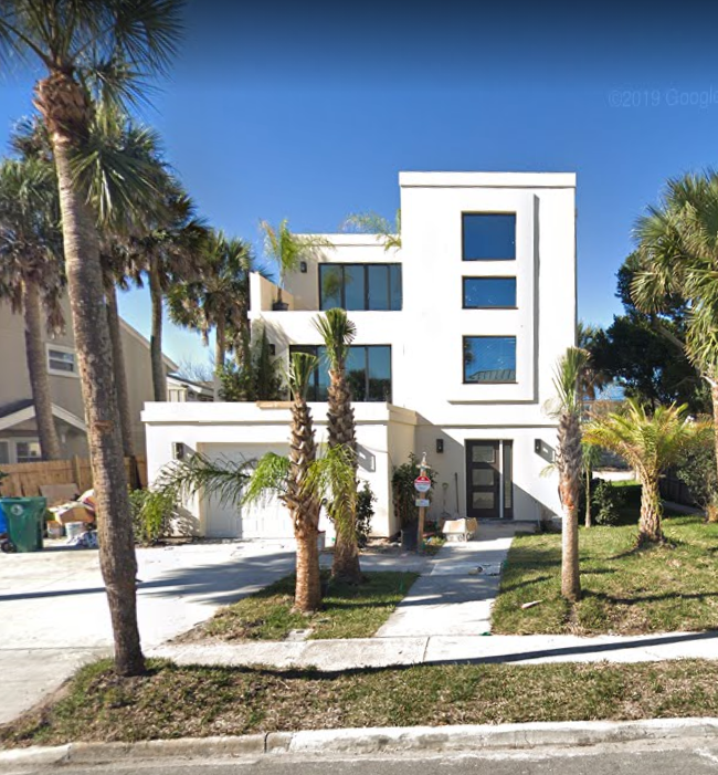 Modern white multi-story house with large windows, surrounded by palm trees and a lawn, under a clear blue sky.