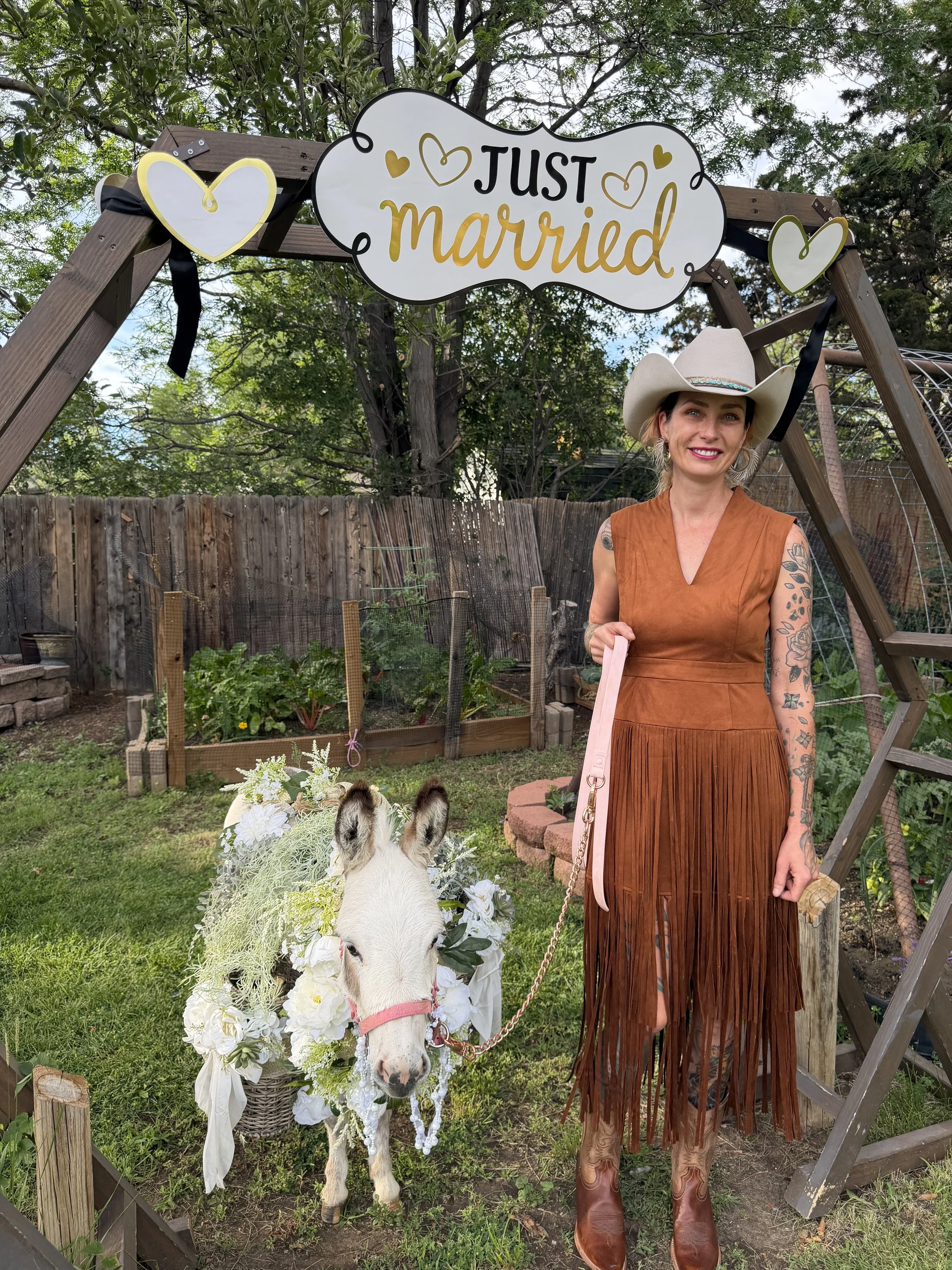 A woman in a fringe dress and cowboy boots standing under a sign that says 'Just Married'. The woman is smiling and holding a leash attached to a white goat decorated with a floral wreath. The background shows a garden with trees, a wooden fence, and a small garden bed.