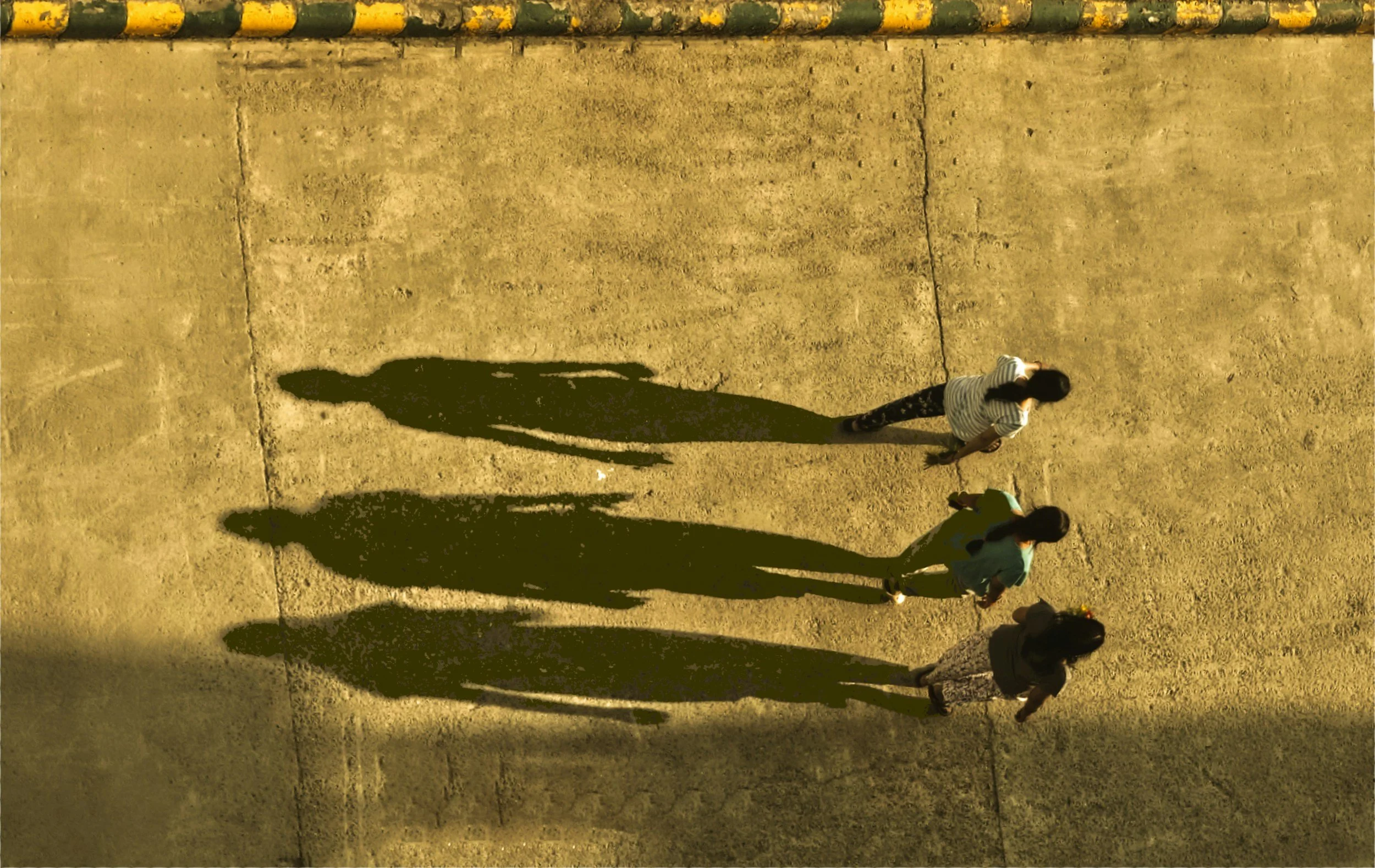 People walking on a sandy ground with their long shadows cast by the sunlight, and a yellow and green striped curb at the top of the image.