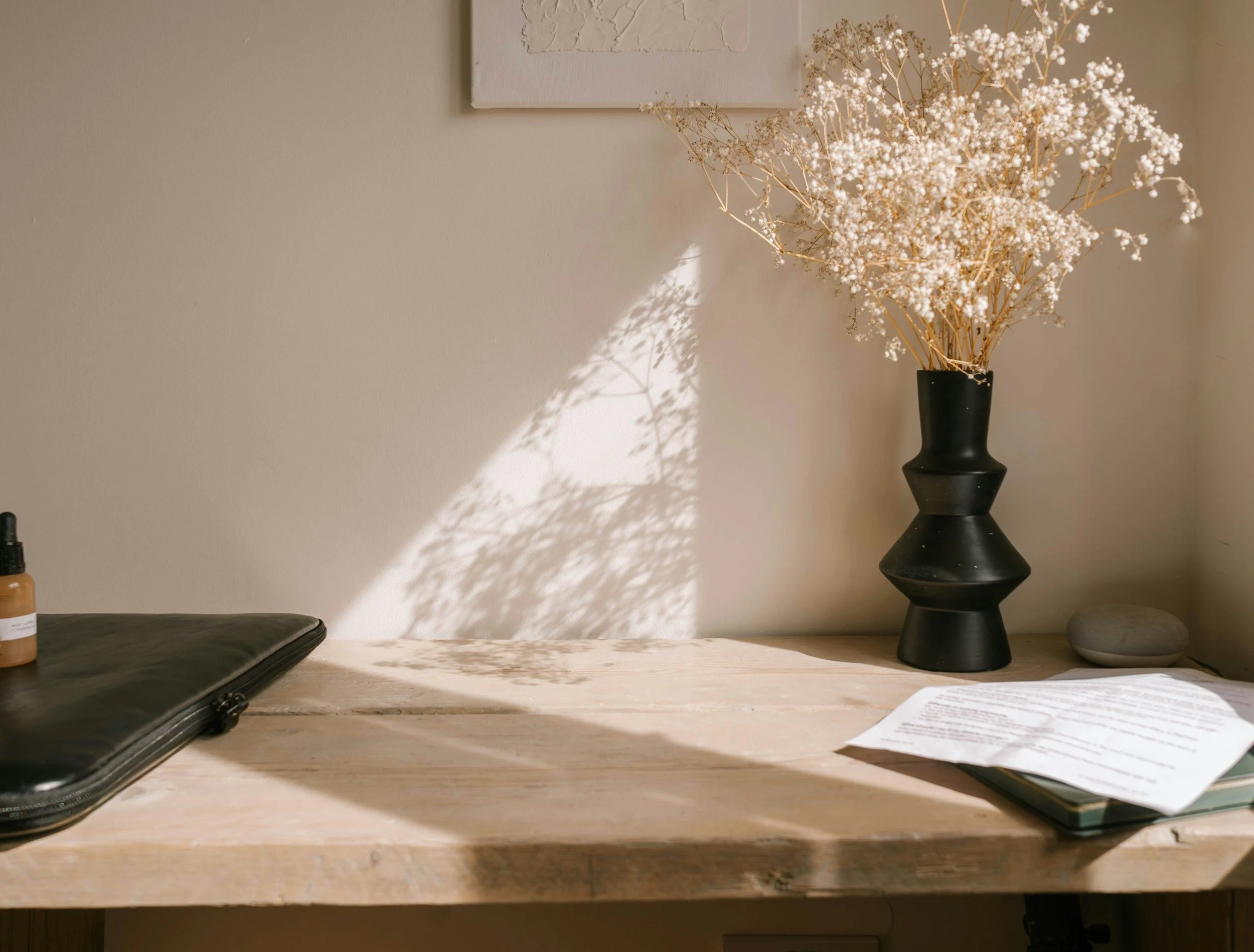 A workspace featuring a black vase with white dried flowers, a laptop, a bottle of supplement, and papers on a light wooden table.