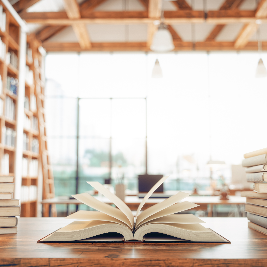 Open book on wooden table in a library or bookstore with large windows and bookshelves in the background.