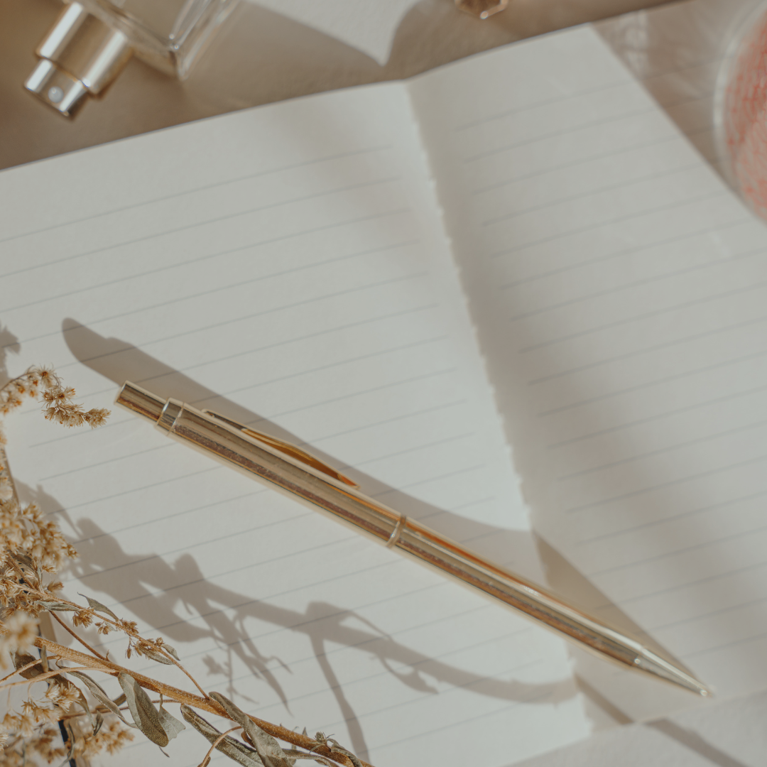Open notebook with lined pages, gold pen, dried flowers, and perfume on a wooden surface with sunlight and shadows.