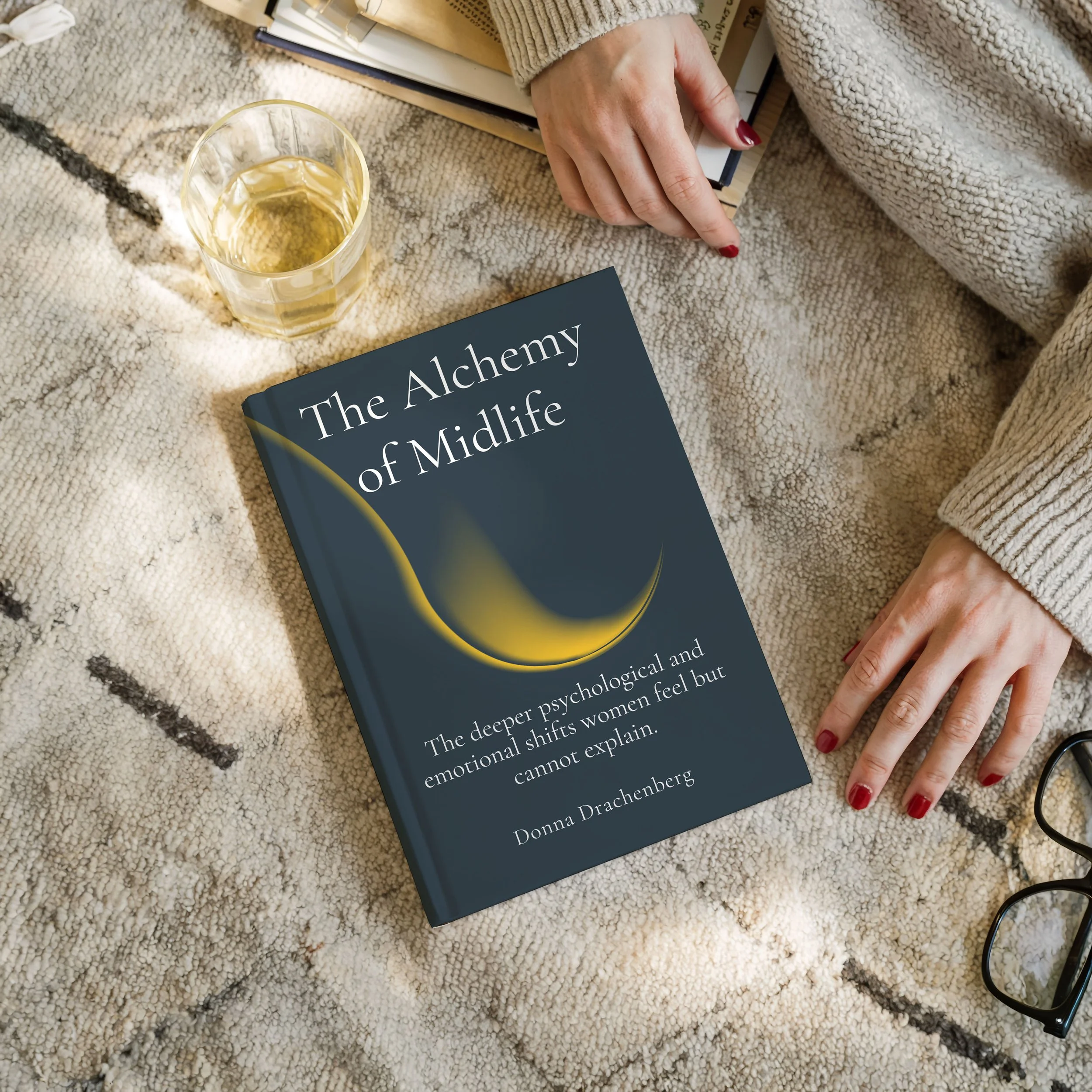 A book titled "The Alchemy of Middle" lies on a textured beige rug. A person's hand with red-painted nails is pointing at the book, and there is a glass of light-colored beverage nearby.