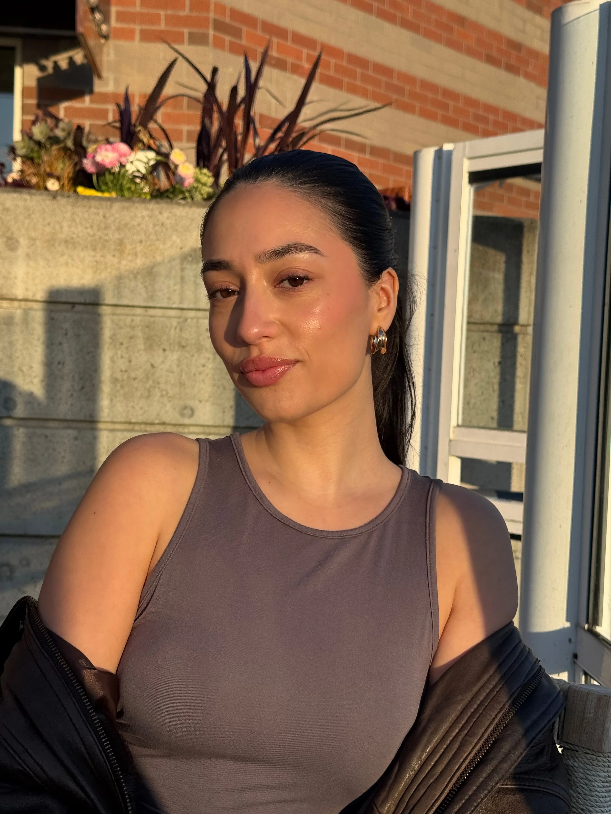 A woman with long dark hair tied back, wearing gold hoop earrings and a sleeveless gray top, taking a selfie outdoors during sunset. She is standing near a concrete wall with flowers and a brick building in the background.