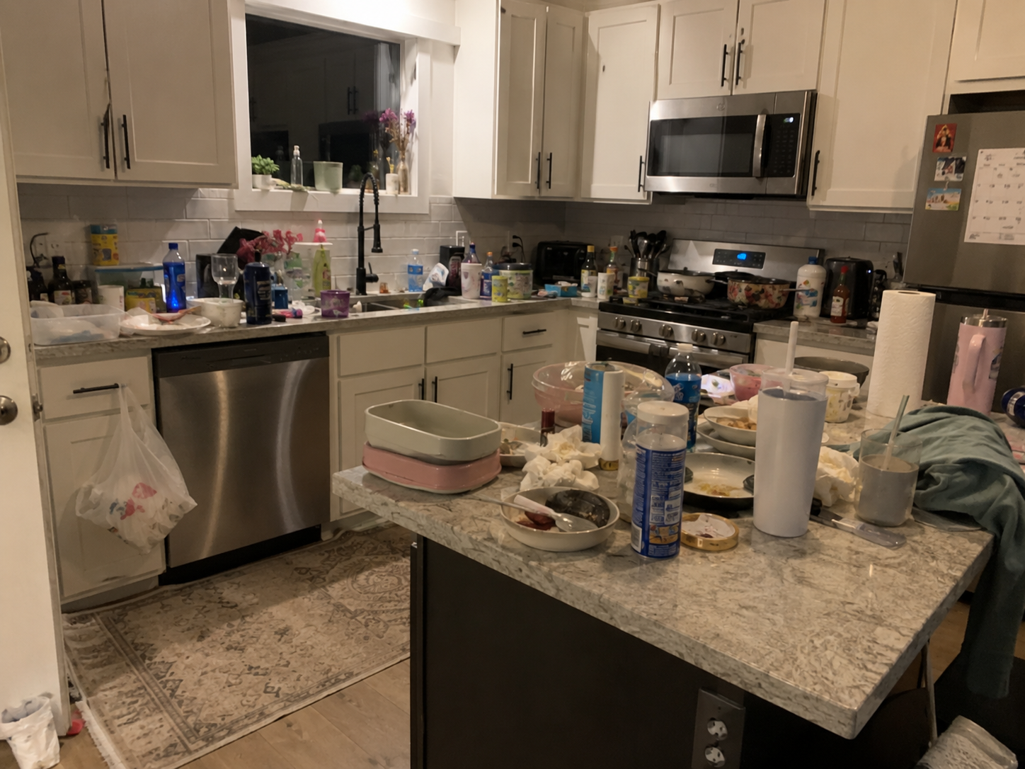 Messy kitchen with cluttered countertops and table, various dishes, drinks, paper towels, and cleaning supplies, white cabinets, stainless steel appliances, and a window above the sink.
