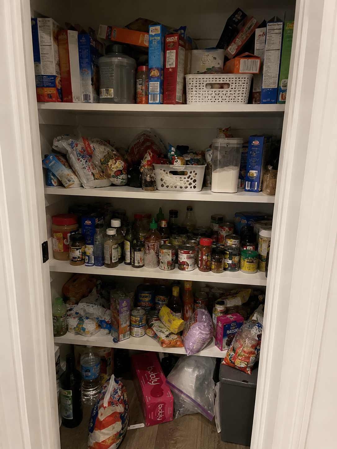 A pantry filled with various food items including canned goods, boxed foods, bottles, jars, plastic bags, and snack packages on white shelves.