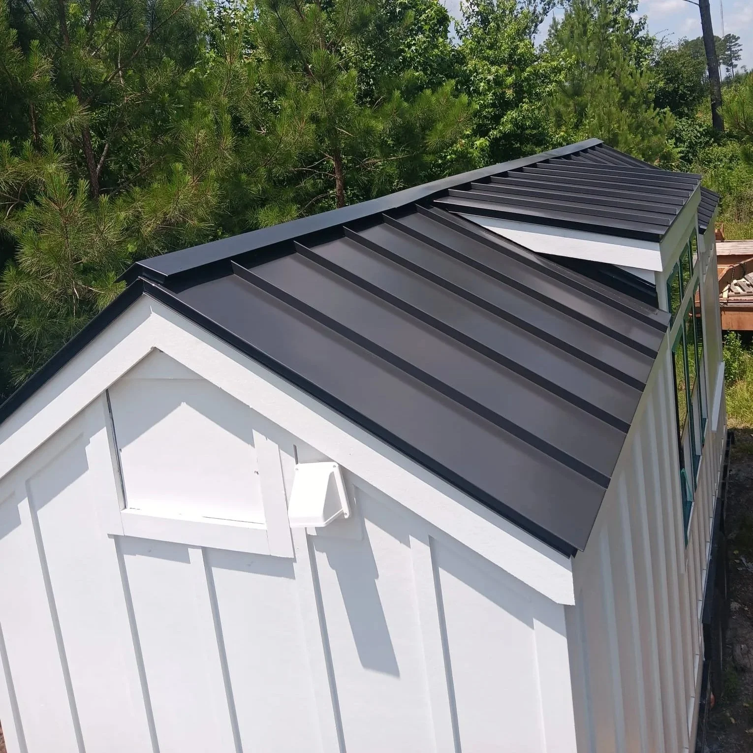 Close-up of the roof of a white house with black metal roofing panels, surrounded by green trees.