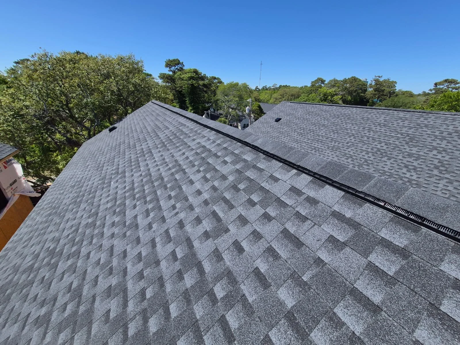 View of a roof with gray asphalt shingles and a black ridge vent, overlooking trees and neighboring houses under a clear blue sky.