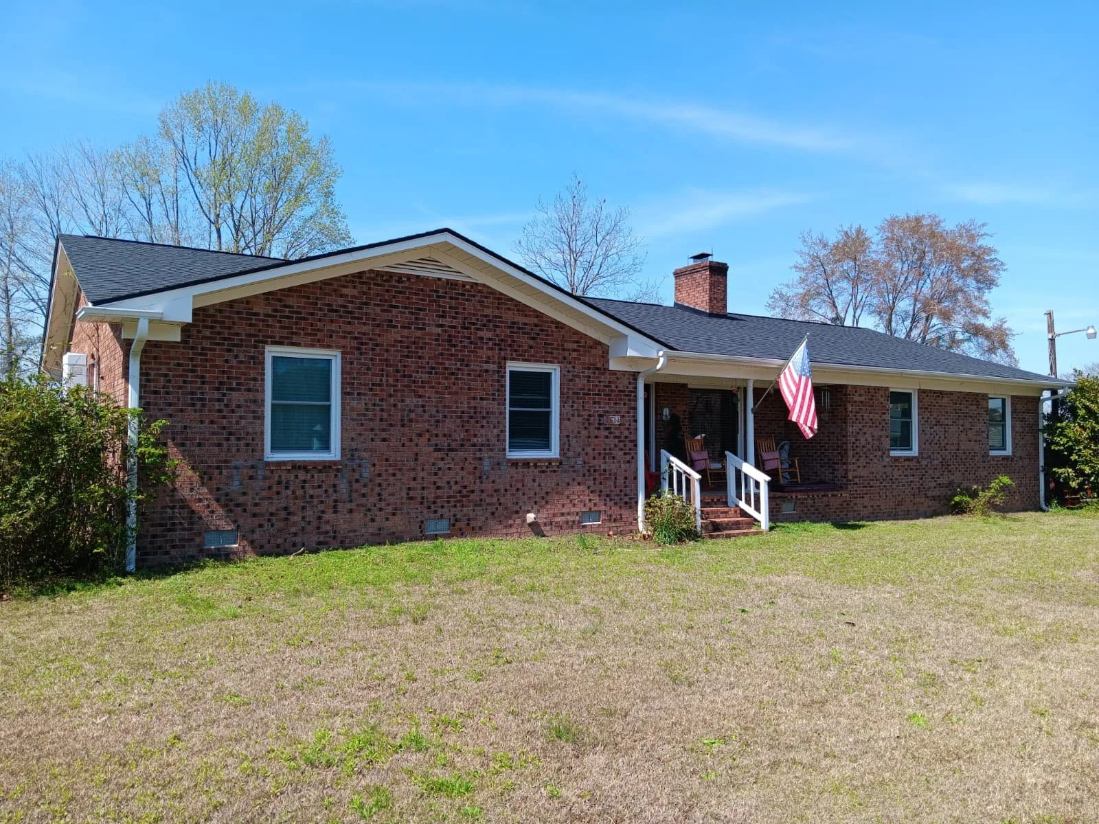 A brick house with three windows, a small porch with porch swings, and an American flag hanging near the front door, set against a clear blue sky with trees in the background.