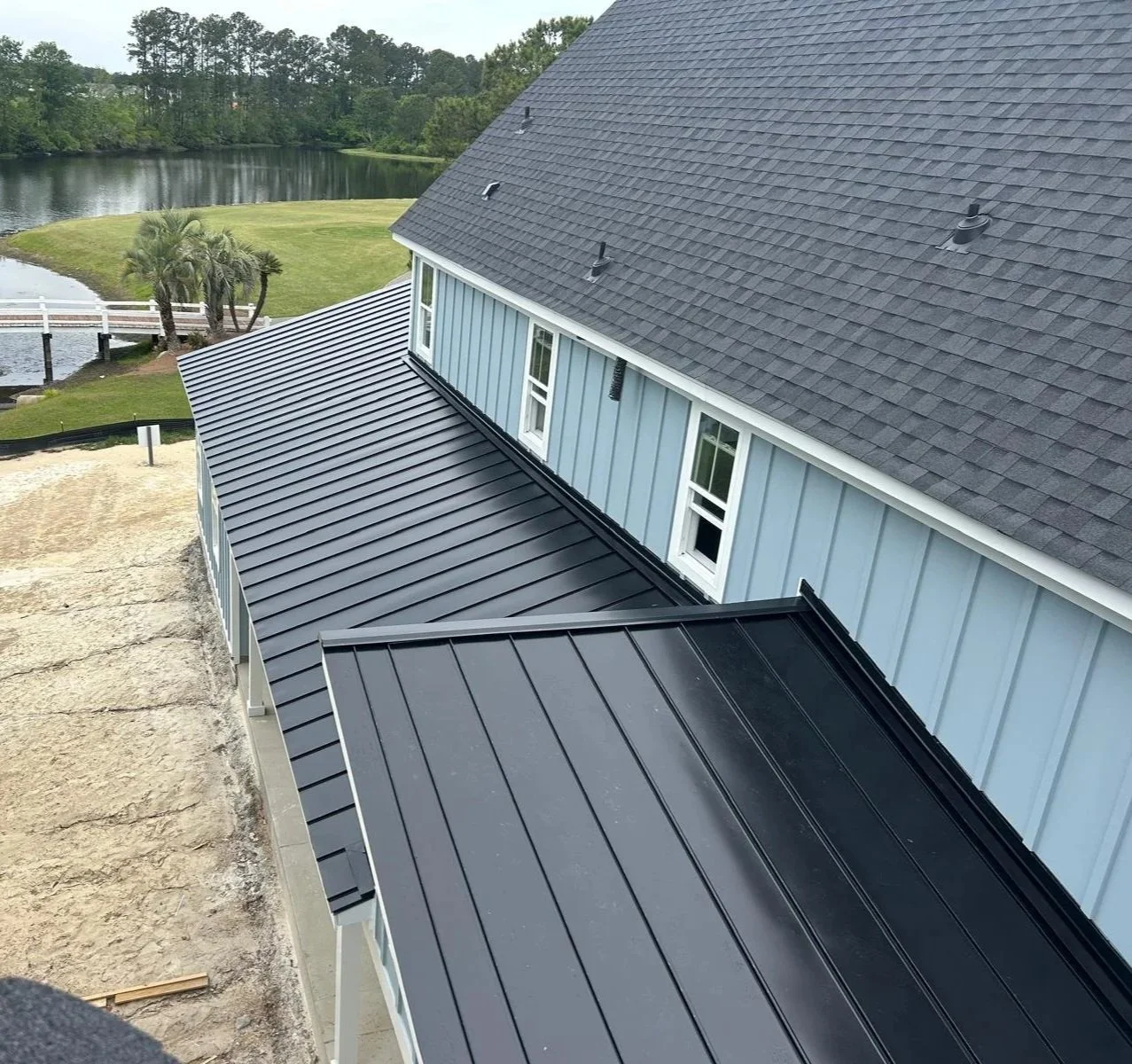 Aerial view of a house with a blue exterior and black metal roofs, overlooking a lake with trees in the background.