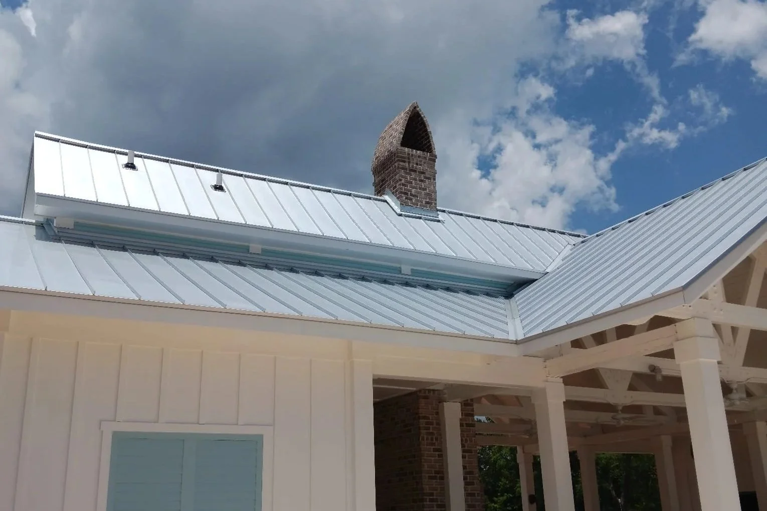 Part of a house with a shiny metal roof, a white exterior wall with a window, and a brick chimney under a cloudy sky.