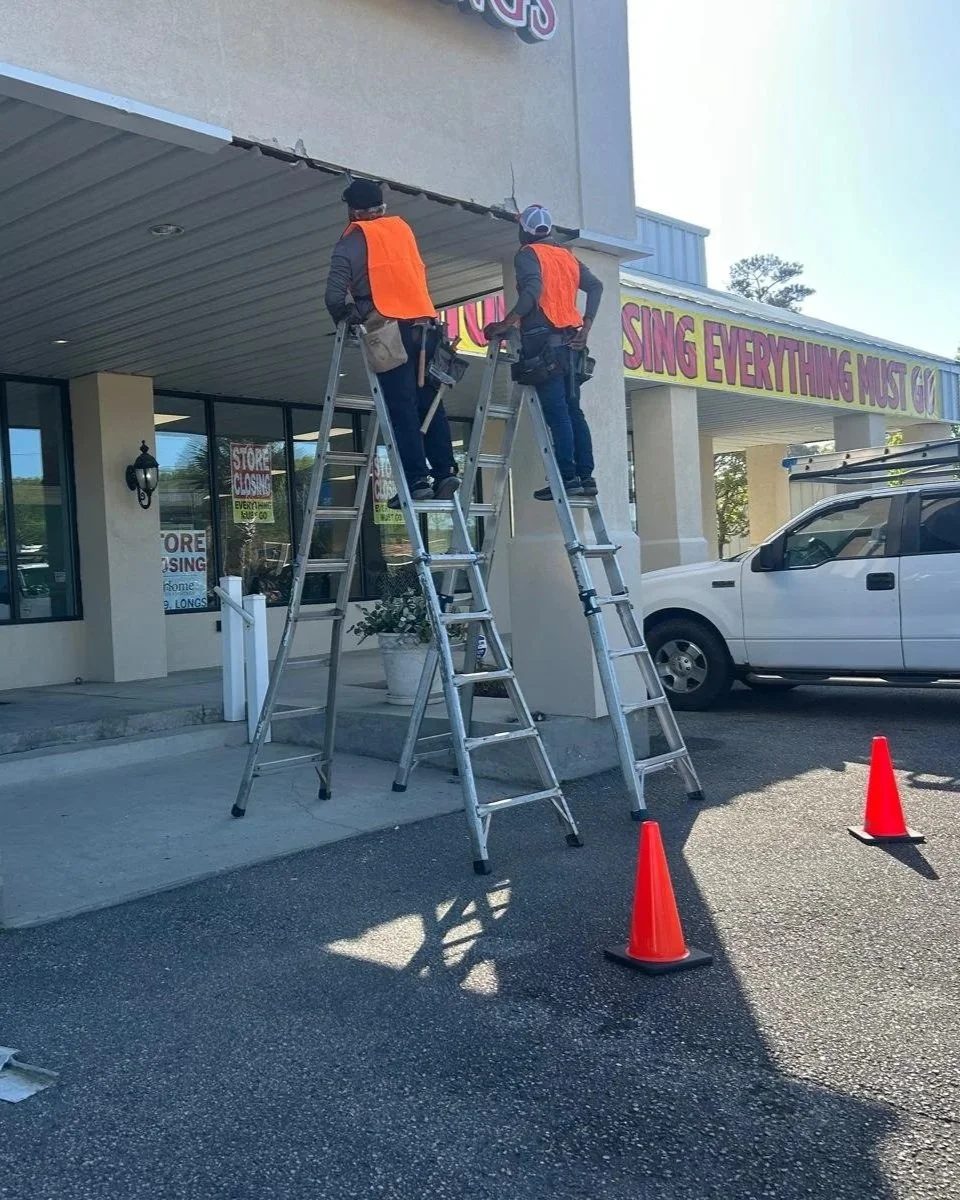 Two workers in orange safety vests on ladders repairing or installing a sign on the building exterior, with traffic cones placed nearby for safety.