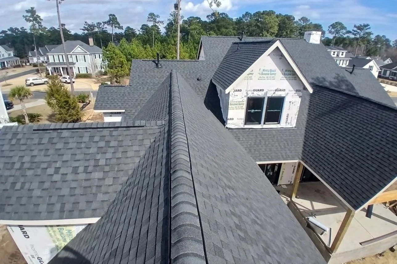 Aerial view of a house under construction with gray asphalt shingles on the roof, a dormer window, and unfinished exterior walls, in a neighborhood with other completed houses and trees in the background.