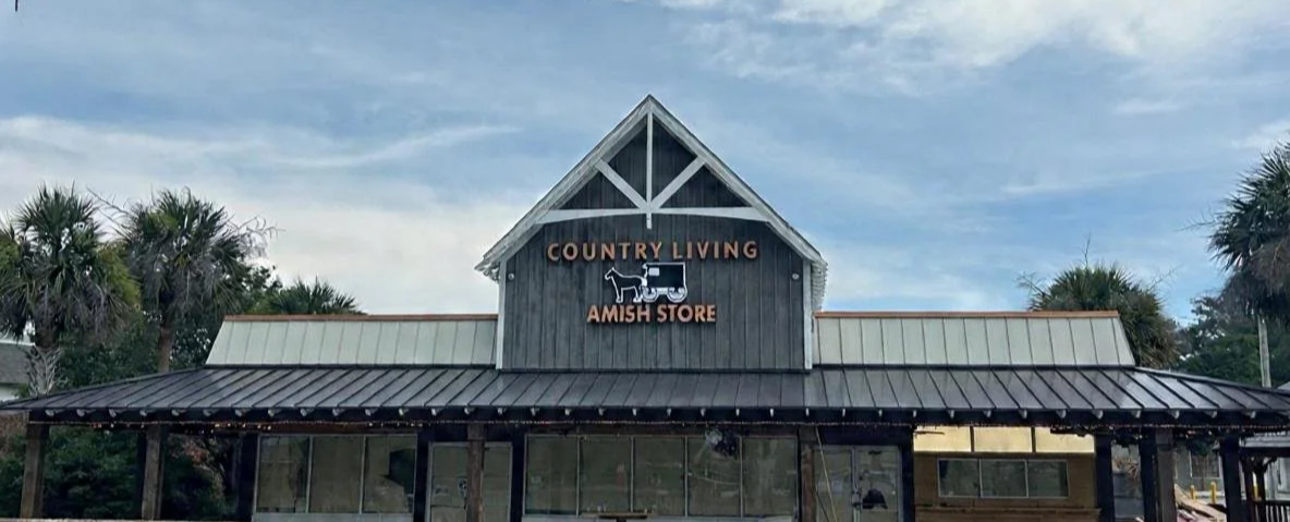 Front view of a rustic country store with a sign that reads 'Country Living Amish Store' and a graphic of a horse-drawn carriage, set against a partly cloudy sky and surrounded by trees.