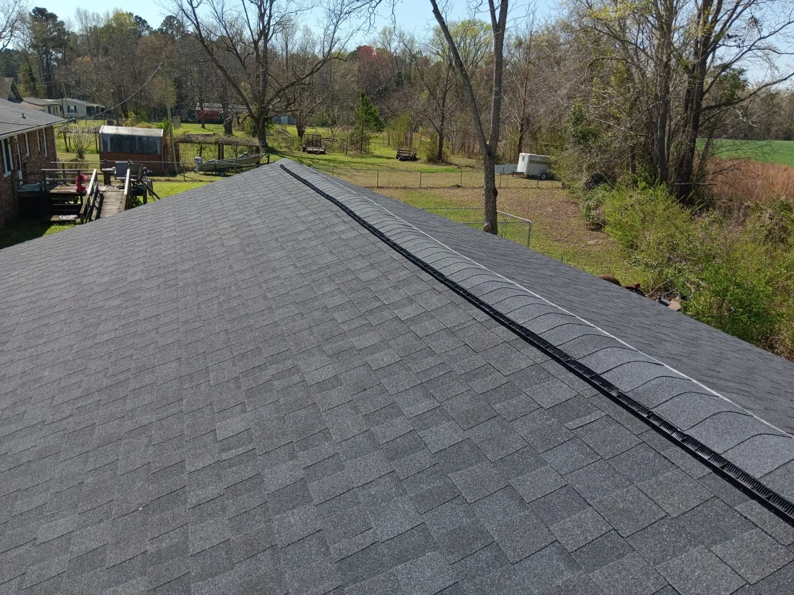 A view of a residential backyard from a rooftop with asphalt shingles, trees, a deck, and outdoor items.