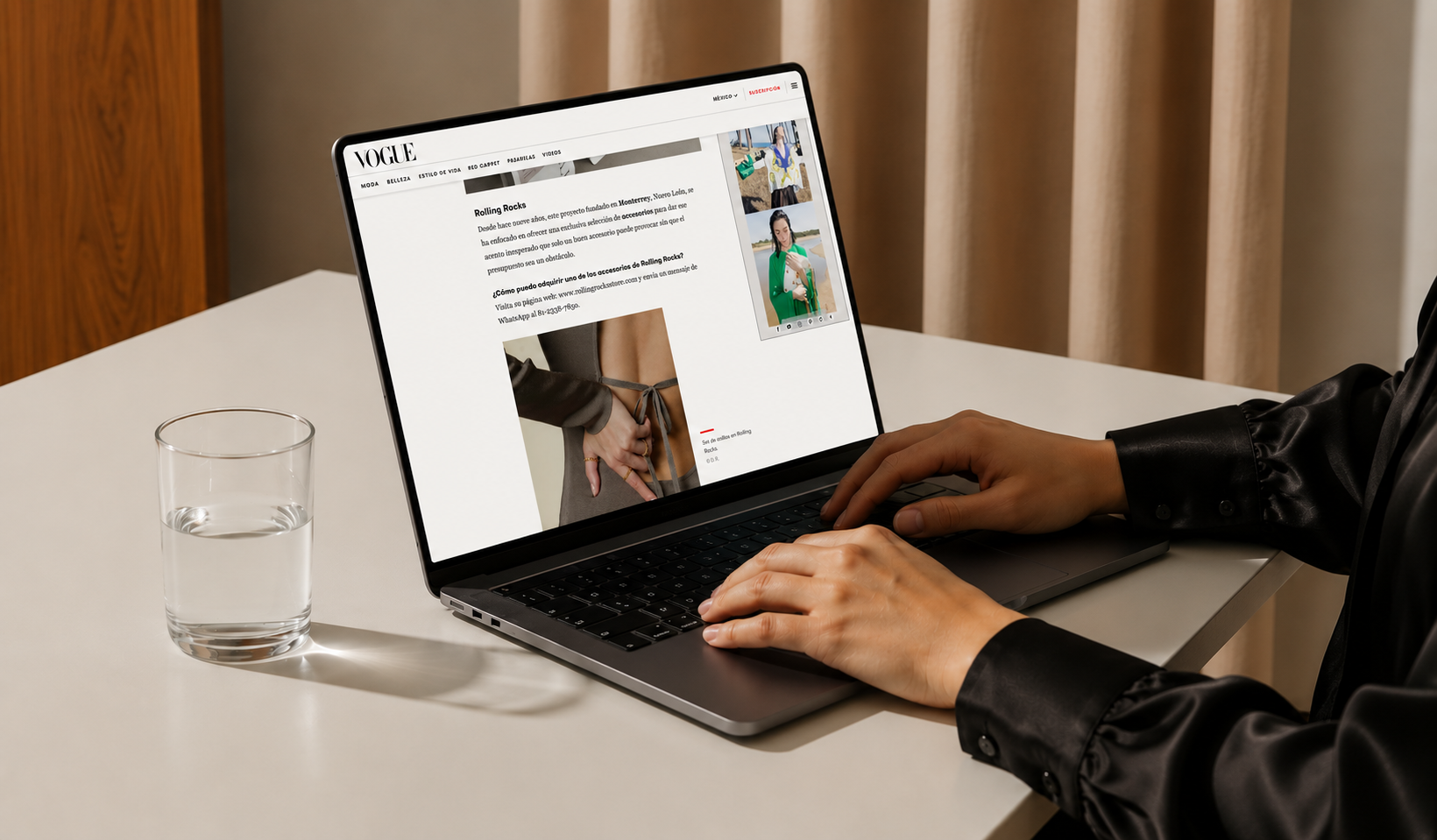 Person in black leather jacket using a laptop on a white desk, reading a fashion article on Vogue website, with a glass of water beside.