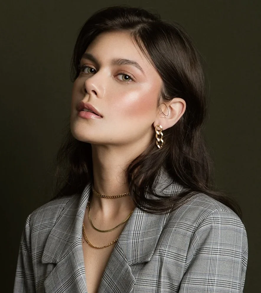 Portrait of a young woman with dark brown hair, green eyes, and light skin, wearing gold jewelry including hoop earrings and layered necklaces, dressed in a gray plaid blazer, against a plain dark background.