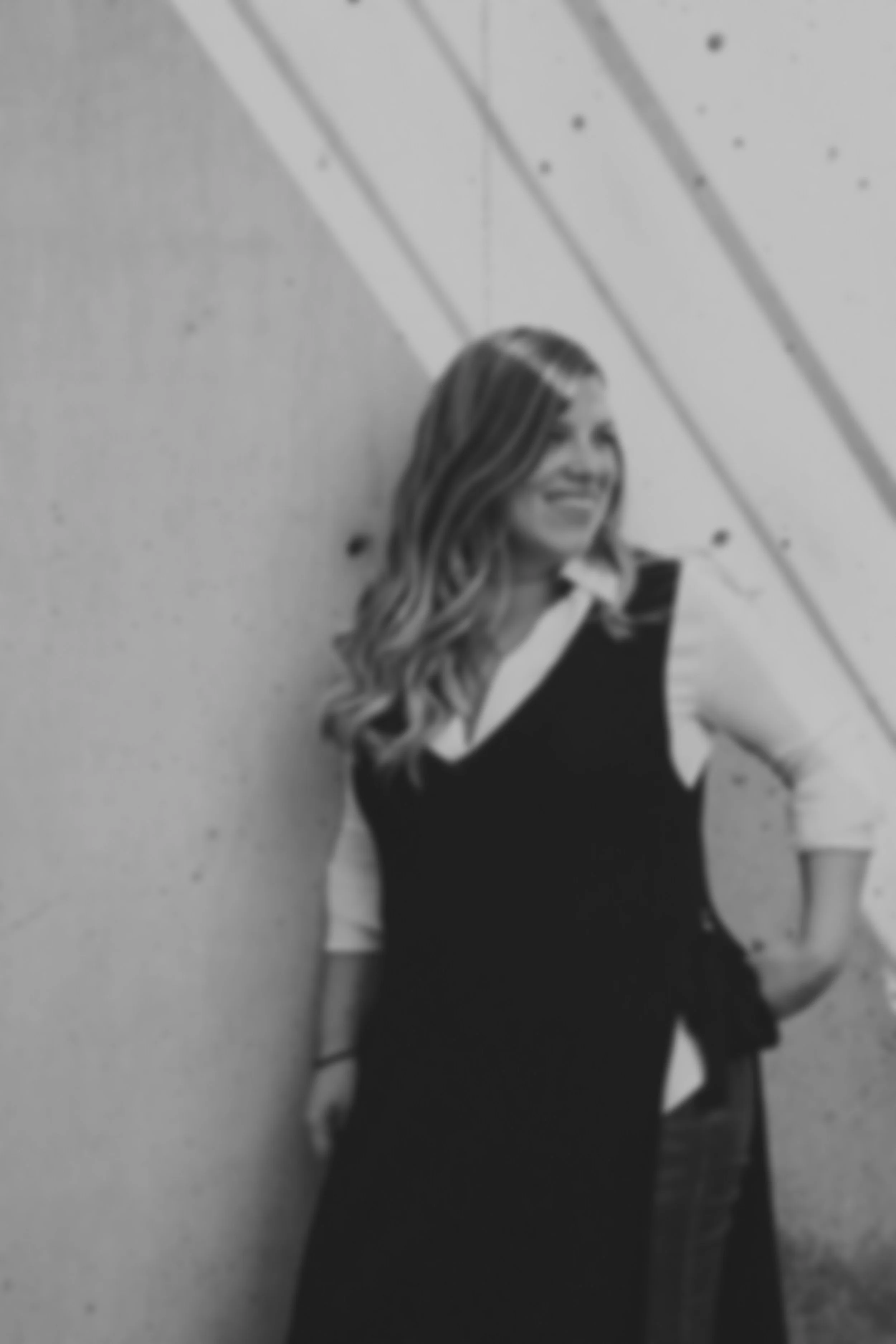 Black and white photo of a smiling woman with long, wavy hair, wearing a black dress with see-through sleeves, standing against a plain wall with diagonal lines.