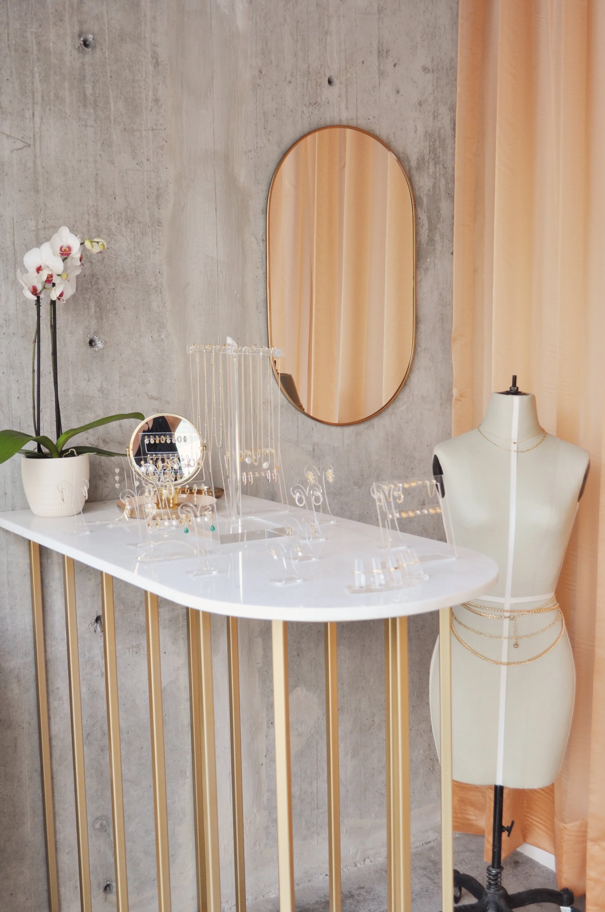 Display of jewelry on a white mannequin bust, with a mirror, a plant, and jewelry stands on a white table, set against a concrete wall with an arched mirror and beige curtains.