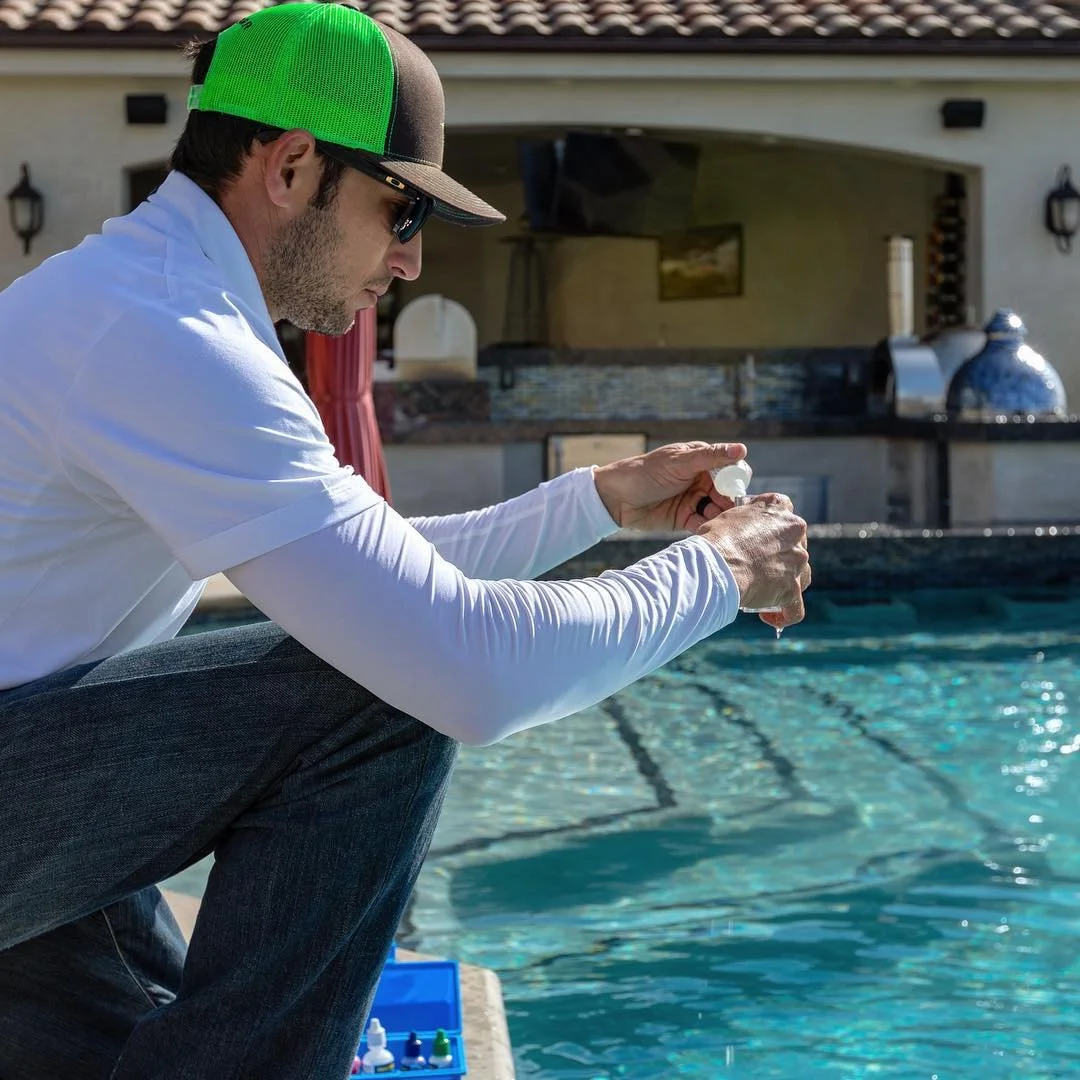 A man kneeling beside a pool, pouring a clear liquid from a small bottle into his hand, wearing sunglasses, a white long-sleeve shirt, and a green and black cap.