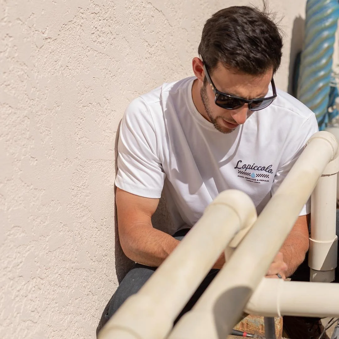 A man with dark hair, wearing sunglasses and a white t-shirt, sitting against a beige textured wall with some PVC pipes in front of him.
