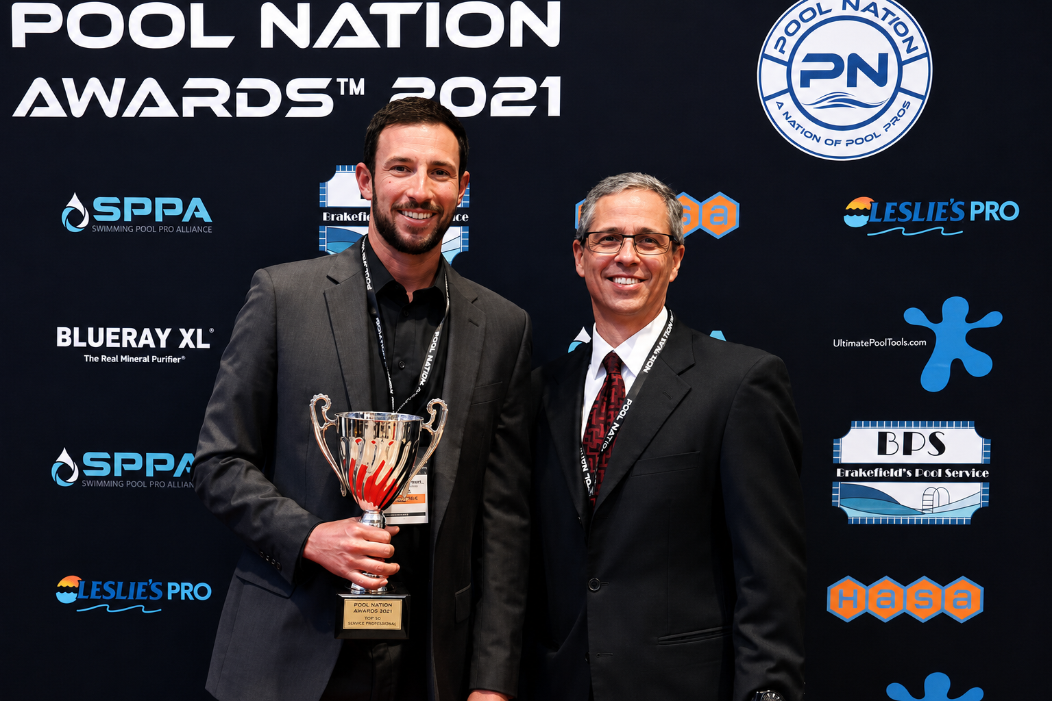 Two men in suits smiling at a pool awards event. The man on the left holds a trophy, and the backdrop displays various pool-related logos and the text 'POOL NATION AWARDS 2021'.