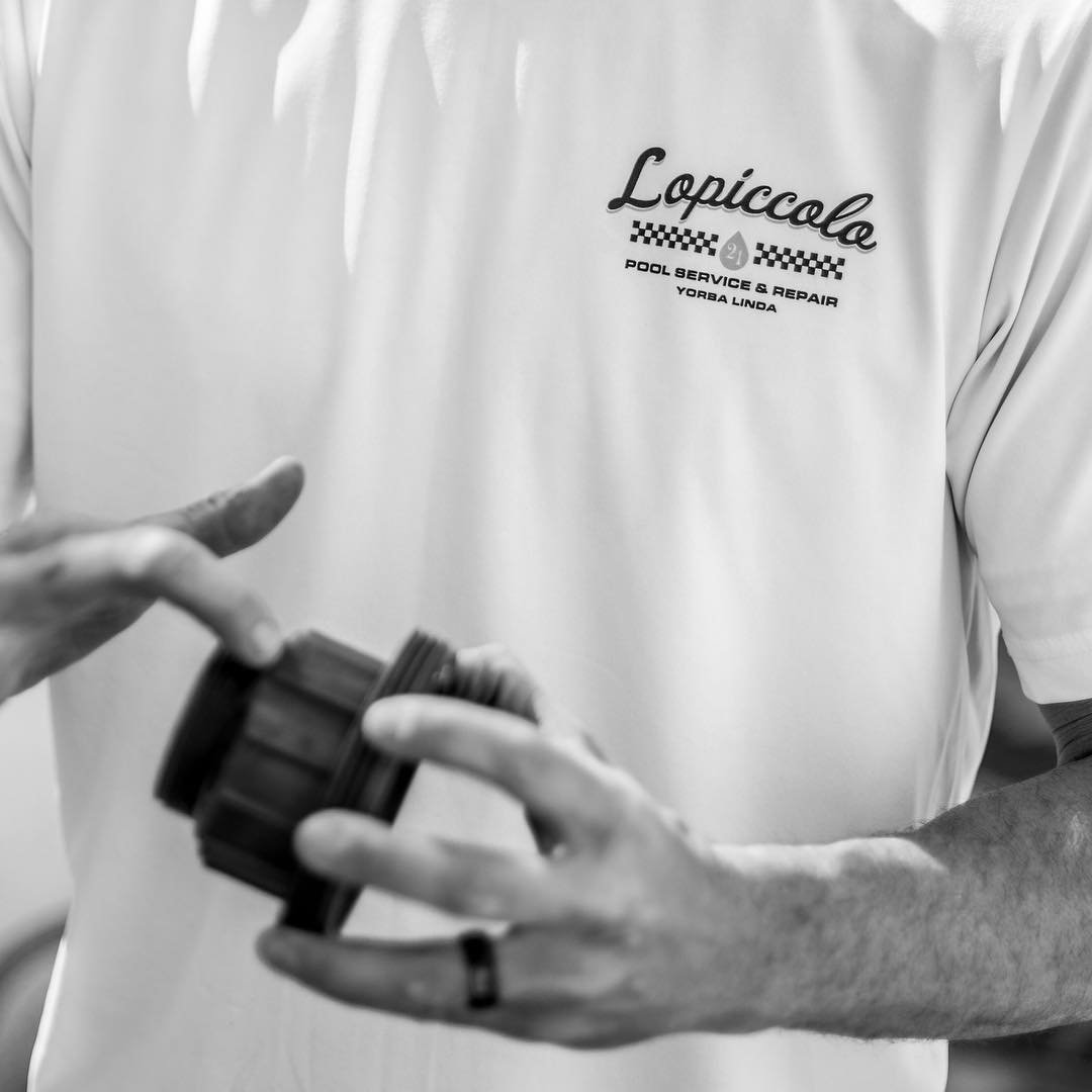 Person holding a black bottle cap, wearing a white polo shirt with the text "Lopiccolo Pool Service & Repair Yorba Linda" logo.