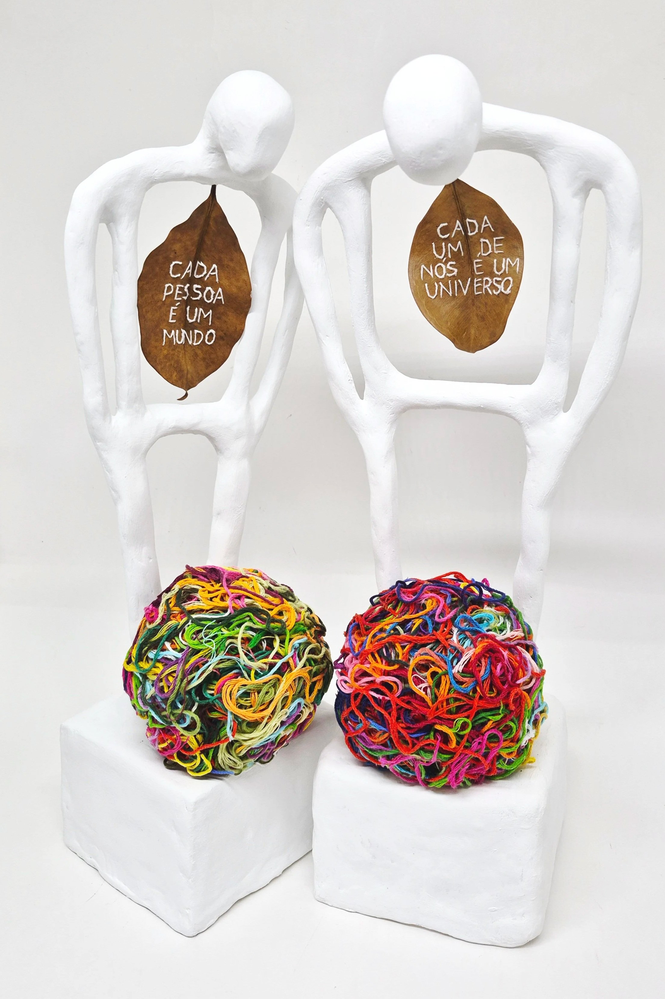 Esculturas de figuras humanas brancas com folhas secas penduradas no pescoço, com palavras escritas nelas em português, e bolas de bolas de lã coloridas na base.