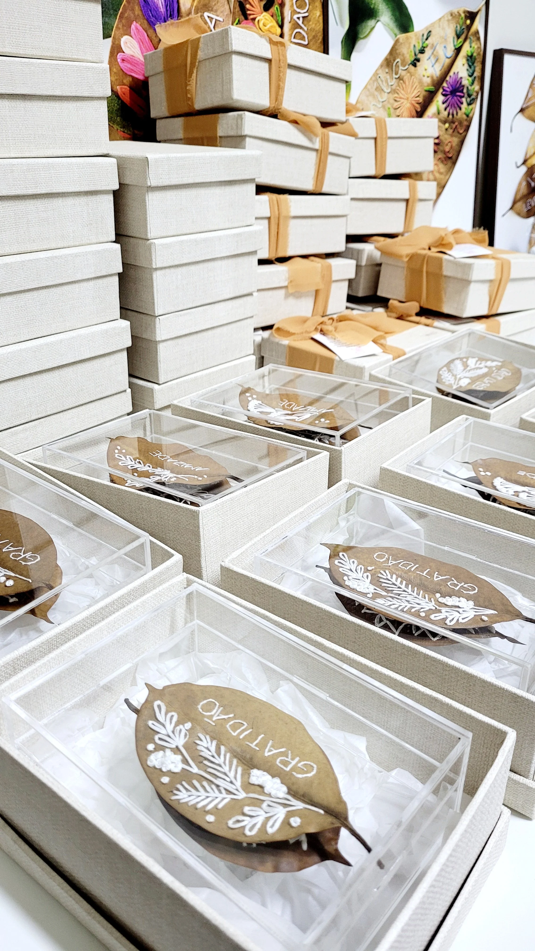 Vários presentes embalados em caixas brancas empilhadas, com laços de fita no topo. Algumas caixas de acrílico com folhas de árvore bordadas a mão com tema de gratidão e folhas ornamentais, com a palavra "Gratidão" escrita.
