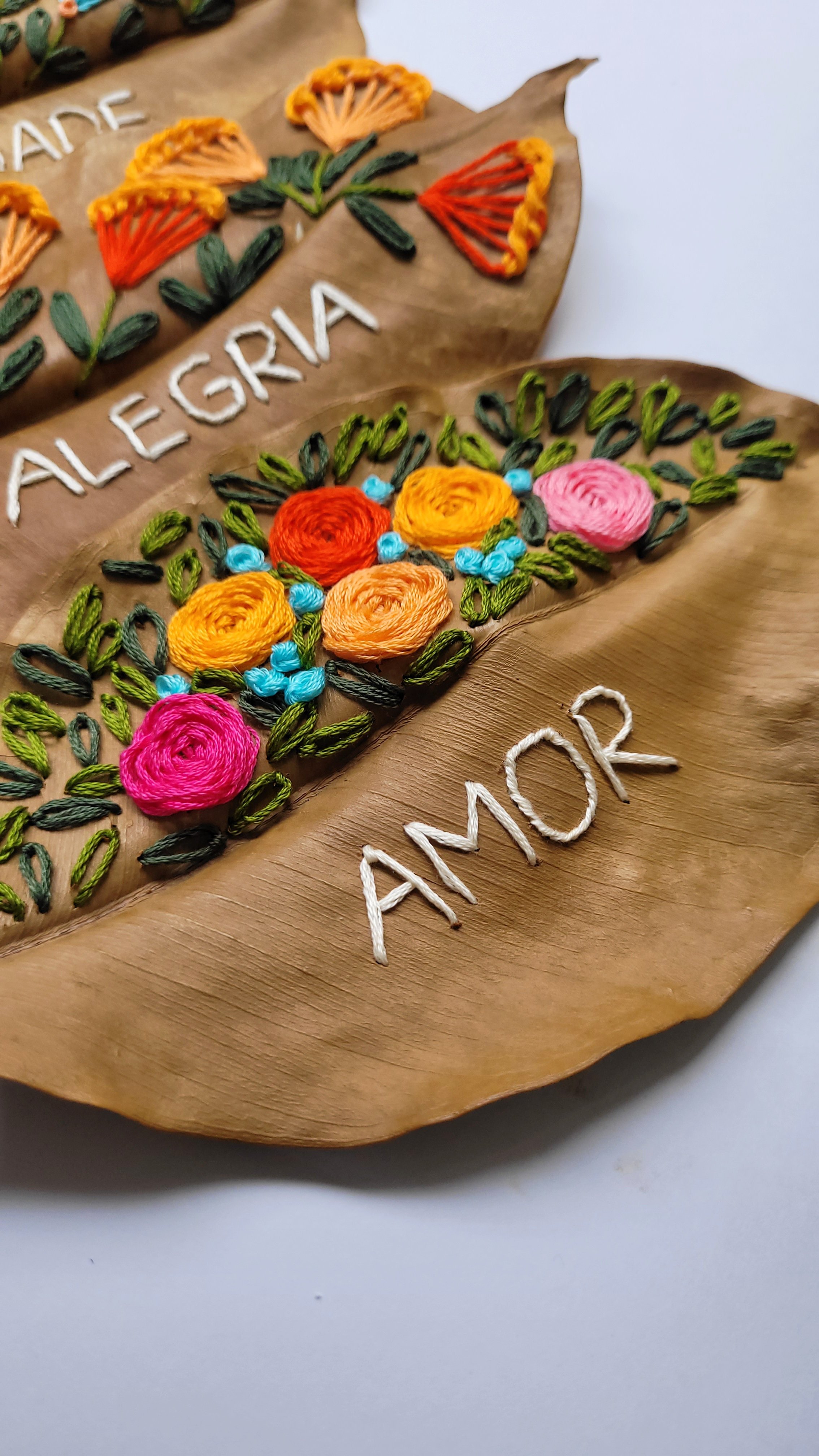 Folhas secas com bordado de flores coloridas feitos com linha de bordado, palavras em português "ALEGRIA" e "AMOR" em pontos brancos.