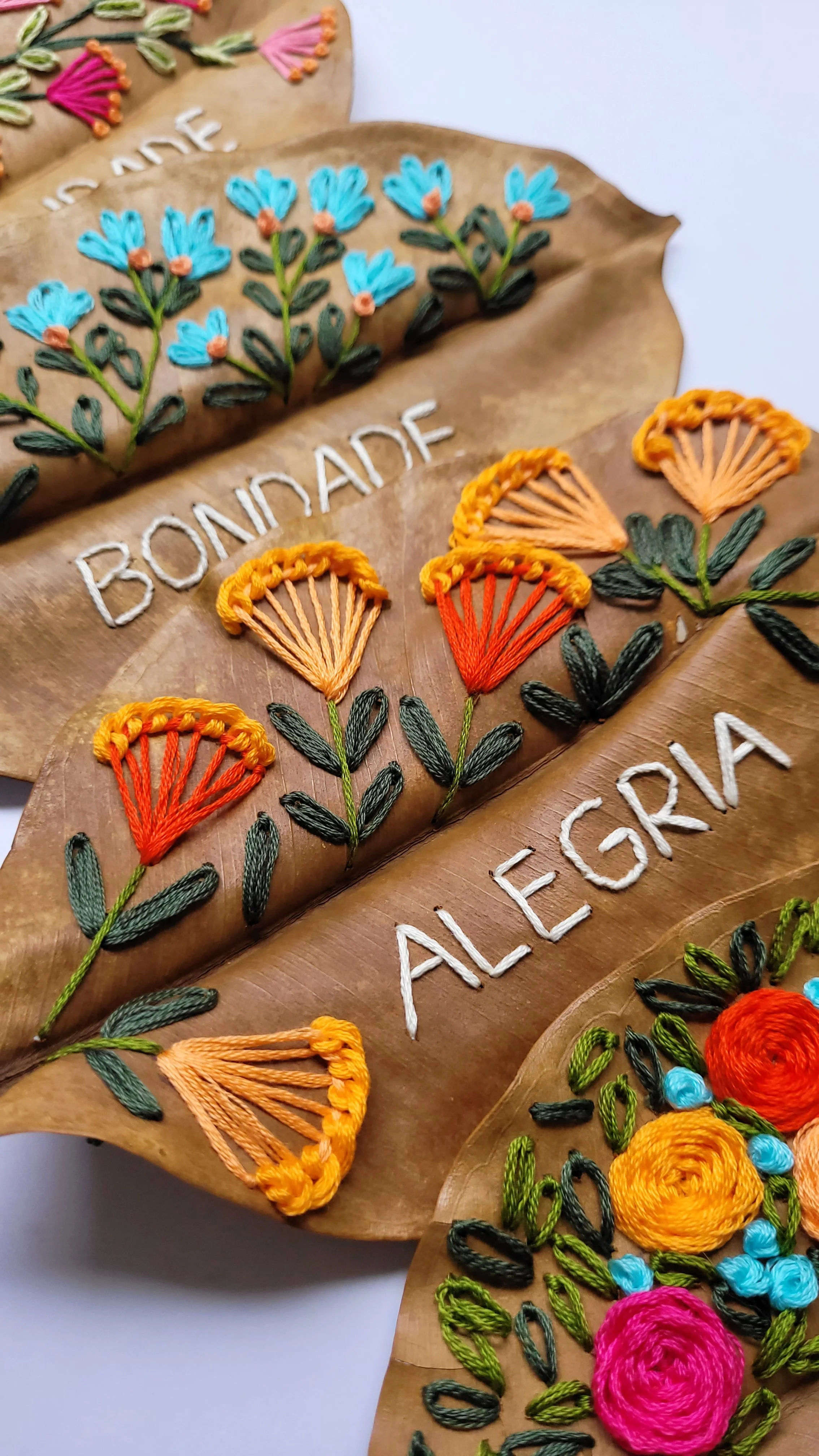 Três folhas naturais com bordado colorido de flores e palavras Bordade e Alegre.