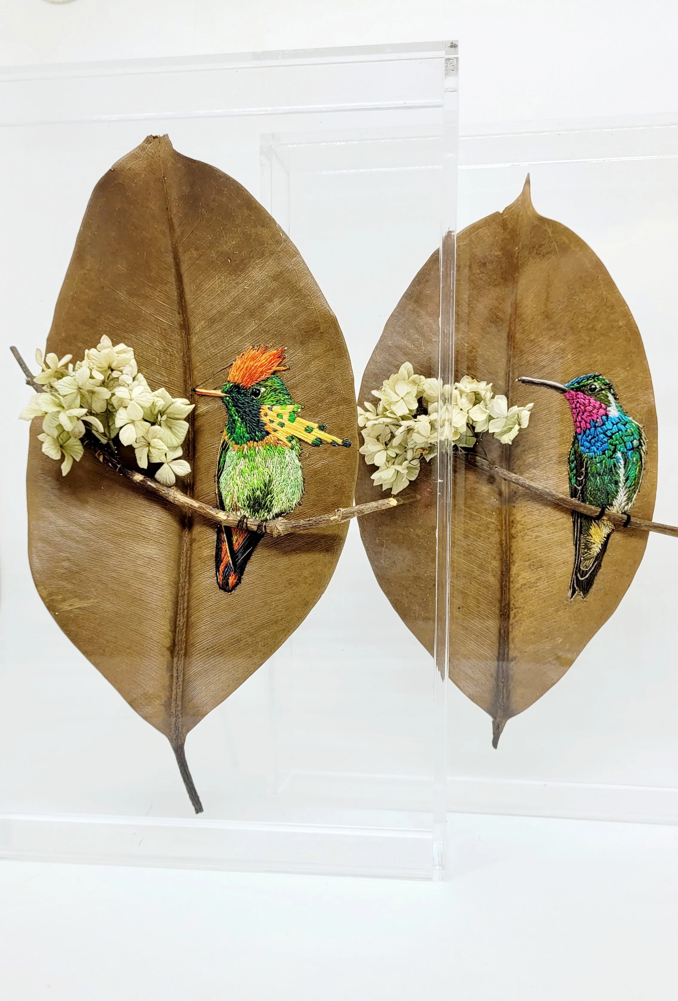 Esculturas de aves feitas com folhas secas, uma de cores vibrantes com penas verdes, amarelas, laranja, e uma com penas azuis, rosa e verdes, colocadas em folhas secas grandes com flores brancas ao lado.