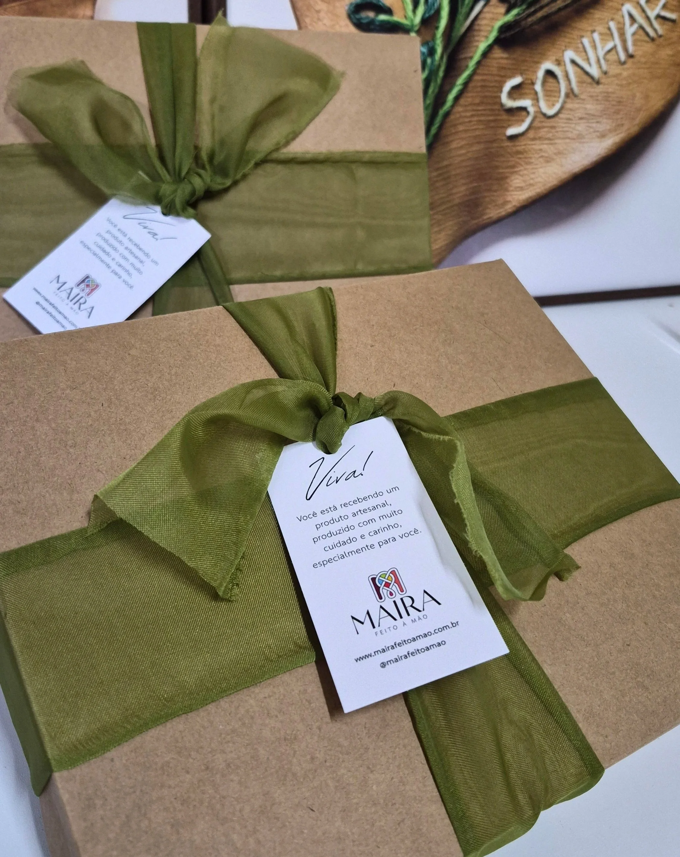 Pacotes de presente embrulhados com papel kraft e acabamento com fita de tecido verde, com etiquetas contendo mensagens de agradecimento e informações da marca Maíra feito à mão, e uma placa de madeira com a palavra "SONHAR" em letras brancas.