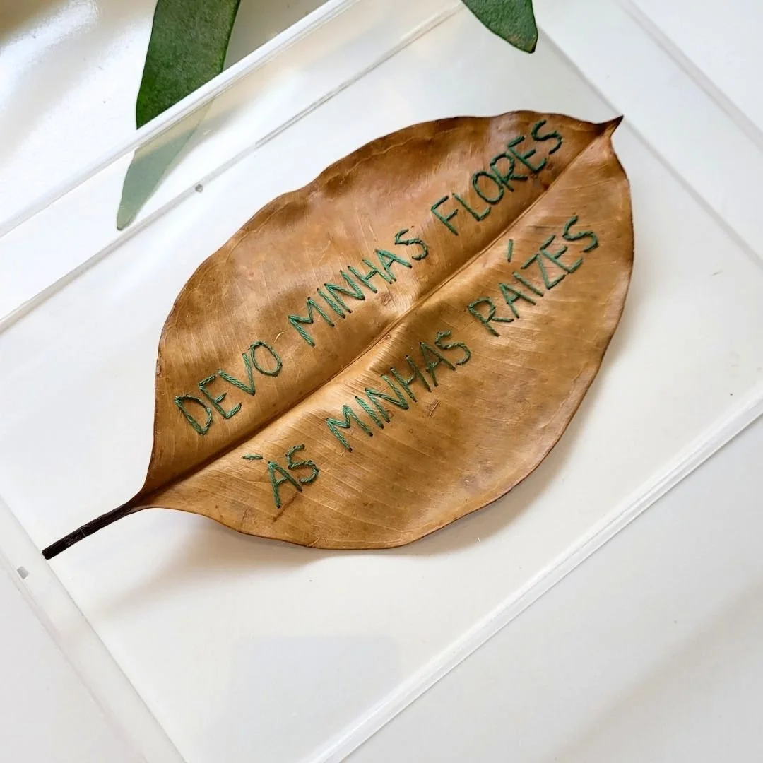 Folha seca com a frase bordada com linha verde: "Devo minhas flores às minhas raízes".