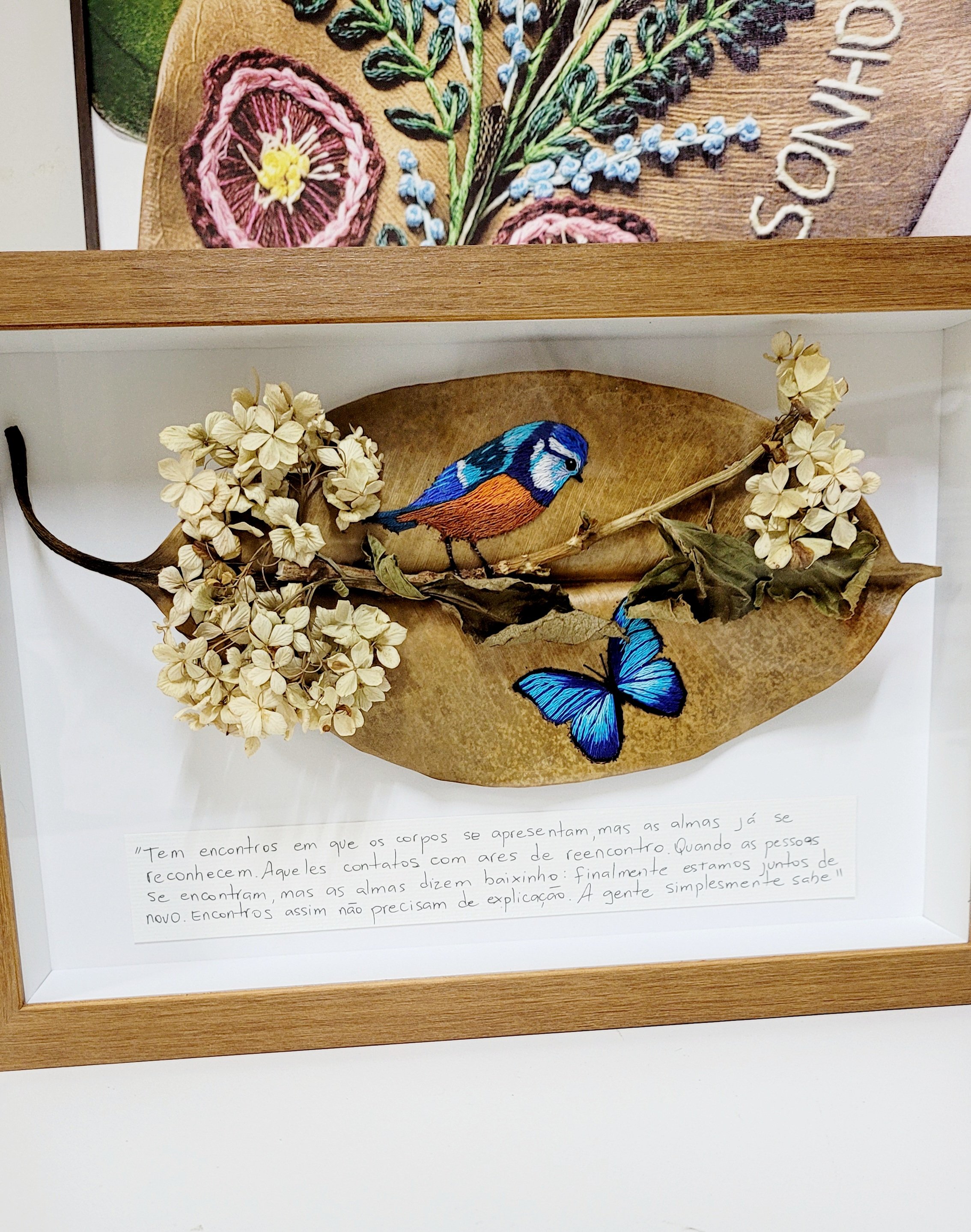 Imagem de uma obra de arte emoldurada, composta por uma peça de madeira com detalhes em bordado que representam elementos da natureza, como flores, um pássaro azul, uma borboleta azul, folhas secas e um trecho de texto em português na parte inferior da moldura.