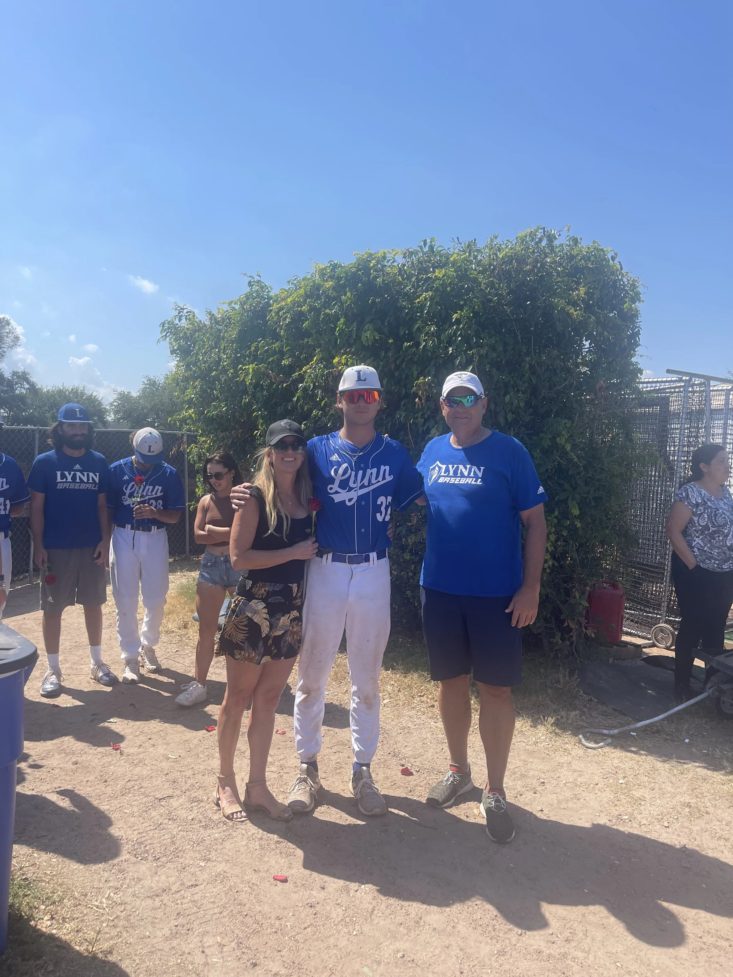 Group of people standing outdoors near a baseball field, some wearing blue and white jerseys with 'Lynn Baseball' on them, with a clear blue sky and green bushes in the background.