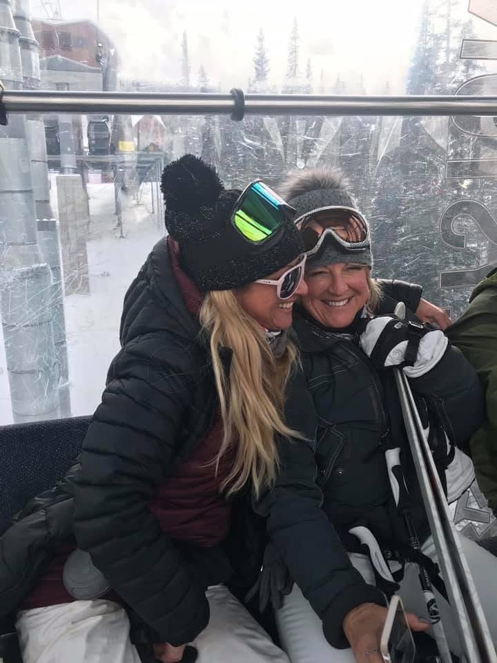 Two women sitting closely together in a ski gondola, smiling and enjoying each other's company with snowy mountain scenery outside.