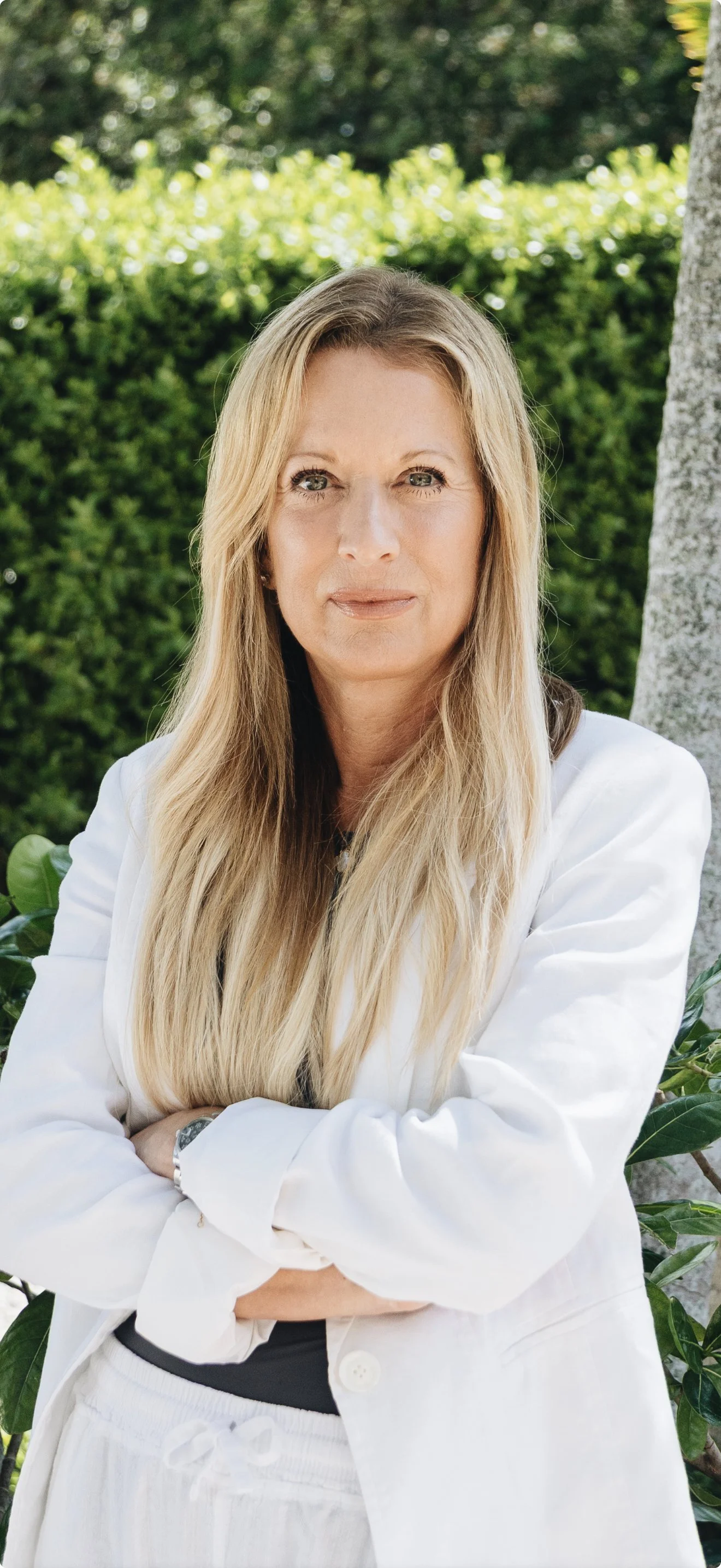 Tracey Potter, a woman with long blonde hair wearing a white blazer and pants, crossing her arms, standing outdoors with green bushes and trees in the background.