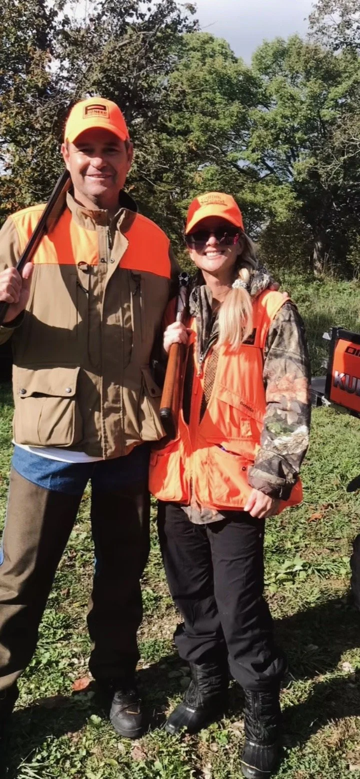 Two people dressed in hunting gear standing outdoors, both wearing orange hats and vests, holding rifles, and smiling at the camera.