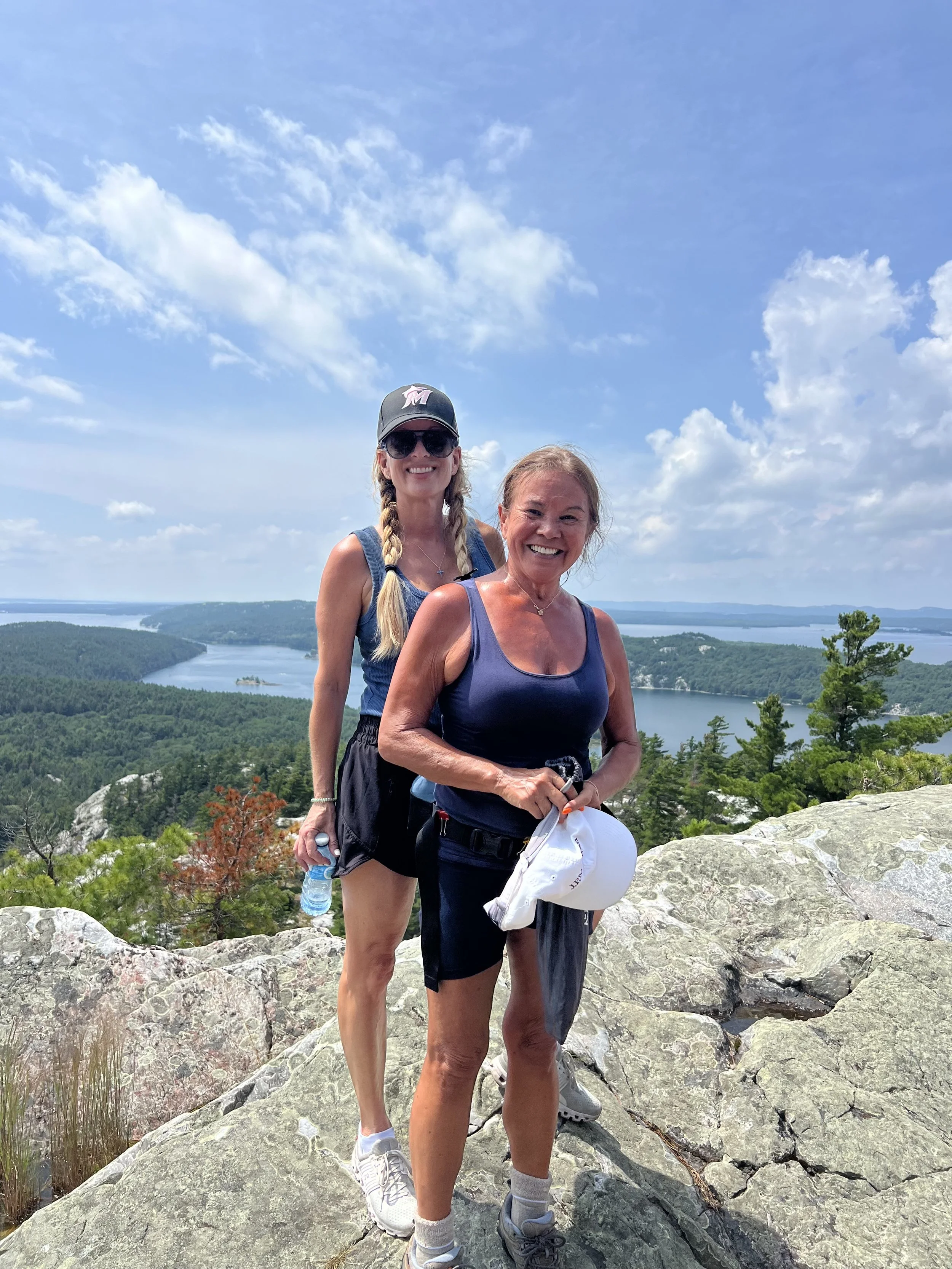 Two women hiking on rocky terrain with a view of a river and forested landscape under a partly cloudy blue sky.