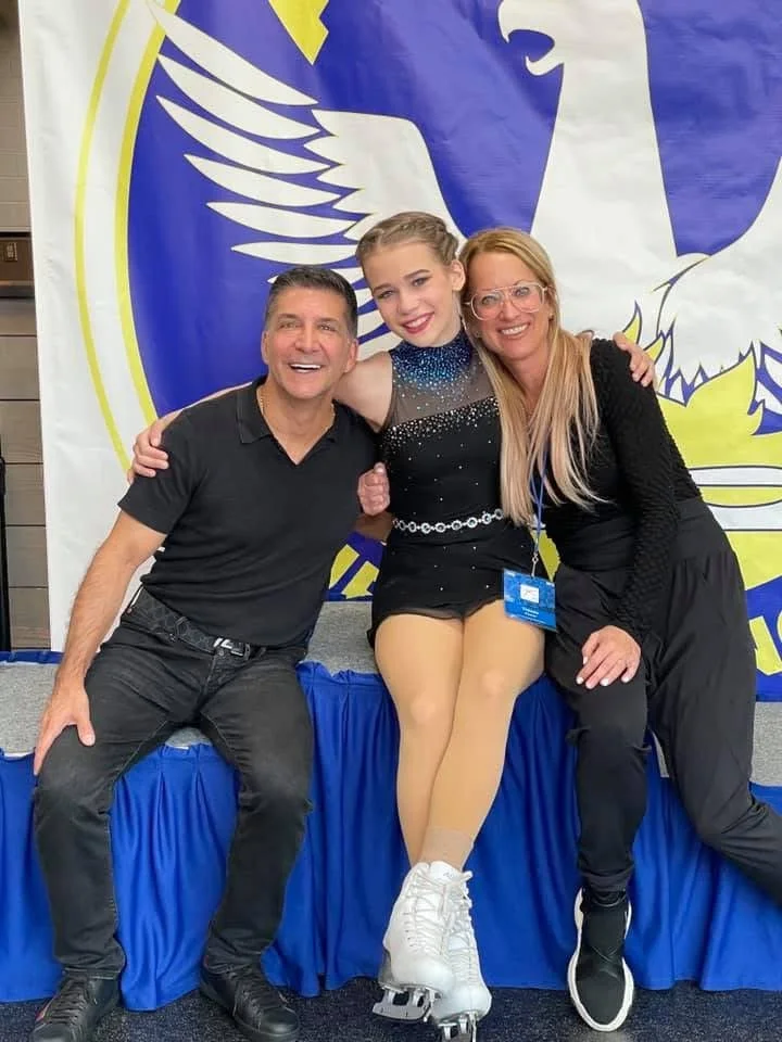 Three people smiling and sitting on a stage in front of a large blue and white banner with an eagle logo. The person in the middle, a young girl in figure skates and a black costume, is hugging a man in a black polo and a woman in black attire.