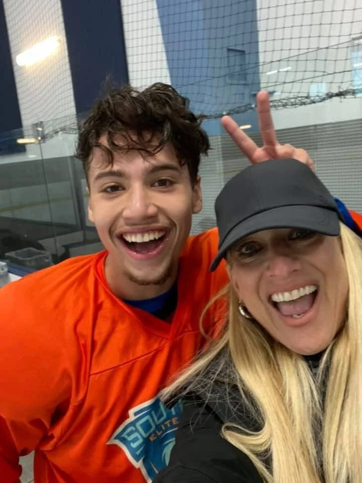 A young man in an orange sports jersey making a peace sign and smiling, taking a selfie with a woman wearing a black cap and a black jacket, both looking happy.