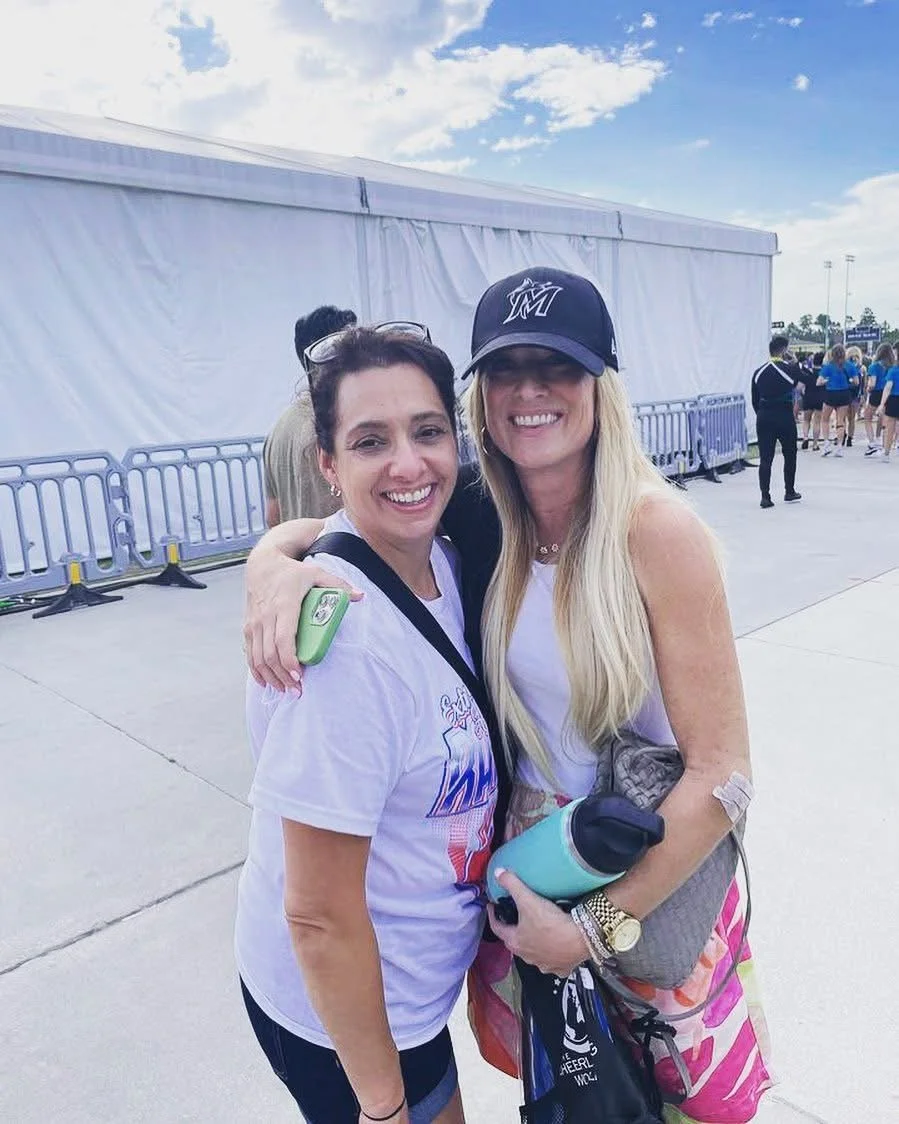 Two women are smiling and hugging each other outdoors, with a large white tent behind them. They are dressed casually, one wearing a Miami Marlins cap and holding a water bottle, the other wearing sunglasses on her head and a white T-shirt with a logo.