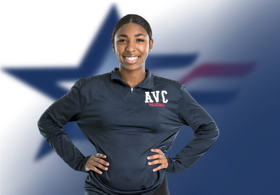A young woman wearing a dark blue AVS Volleyball jacket, standing confidently with hands on hips, smiling, with a blurred American flag in the background.